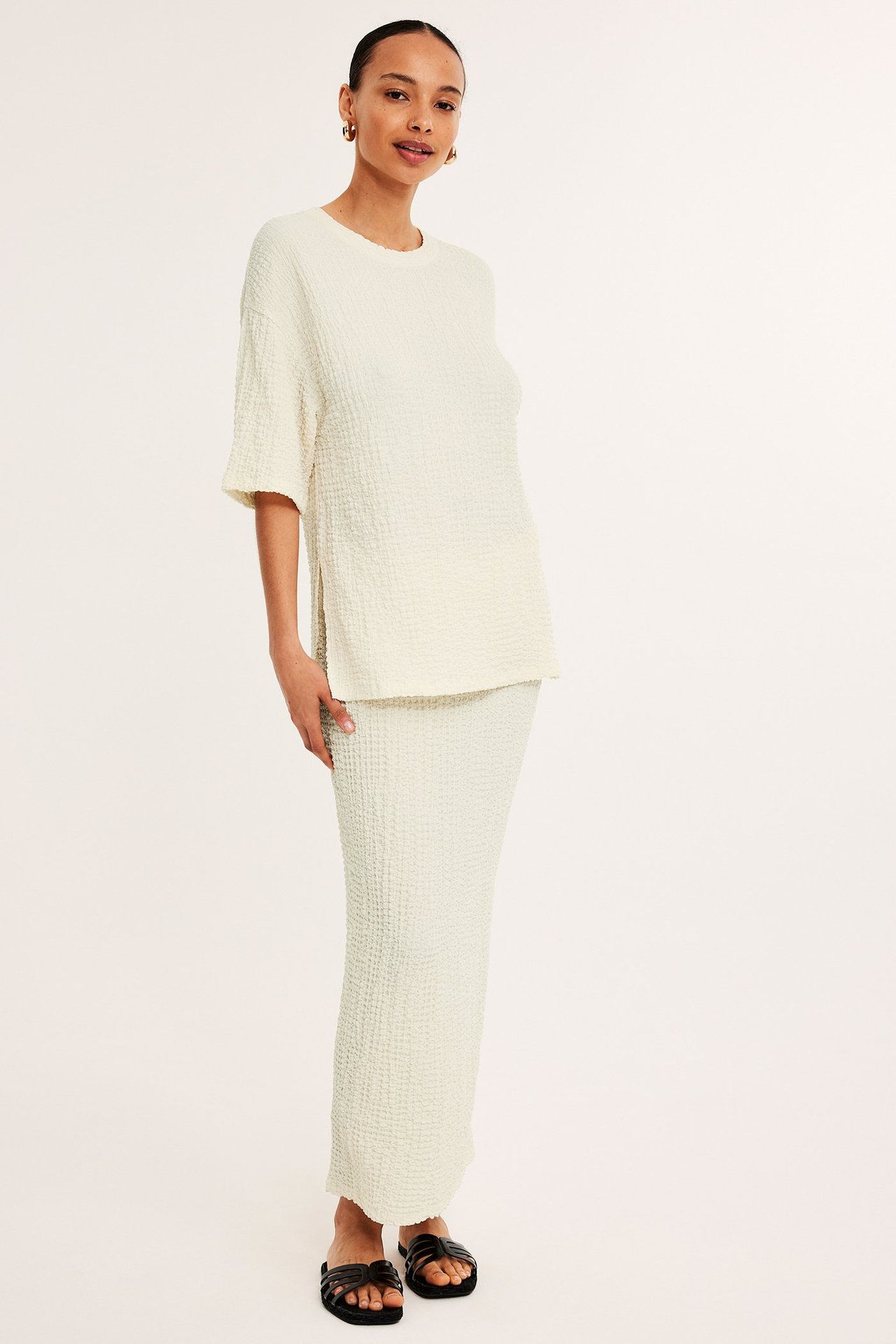 Frontbilde: kvinne i offwhite strukturert kortermet topp, matchende skjørt og svarte sandaler.