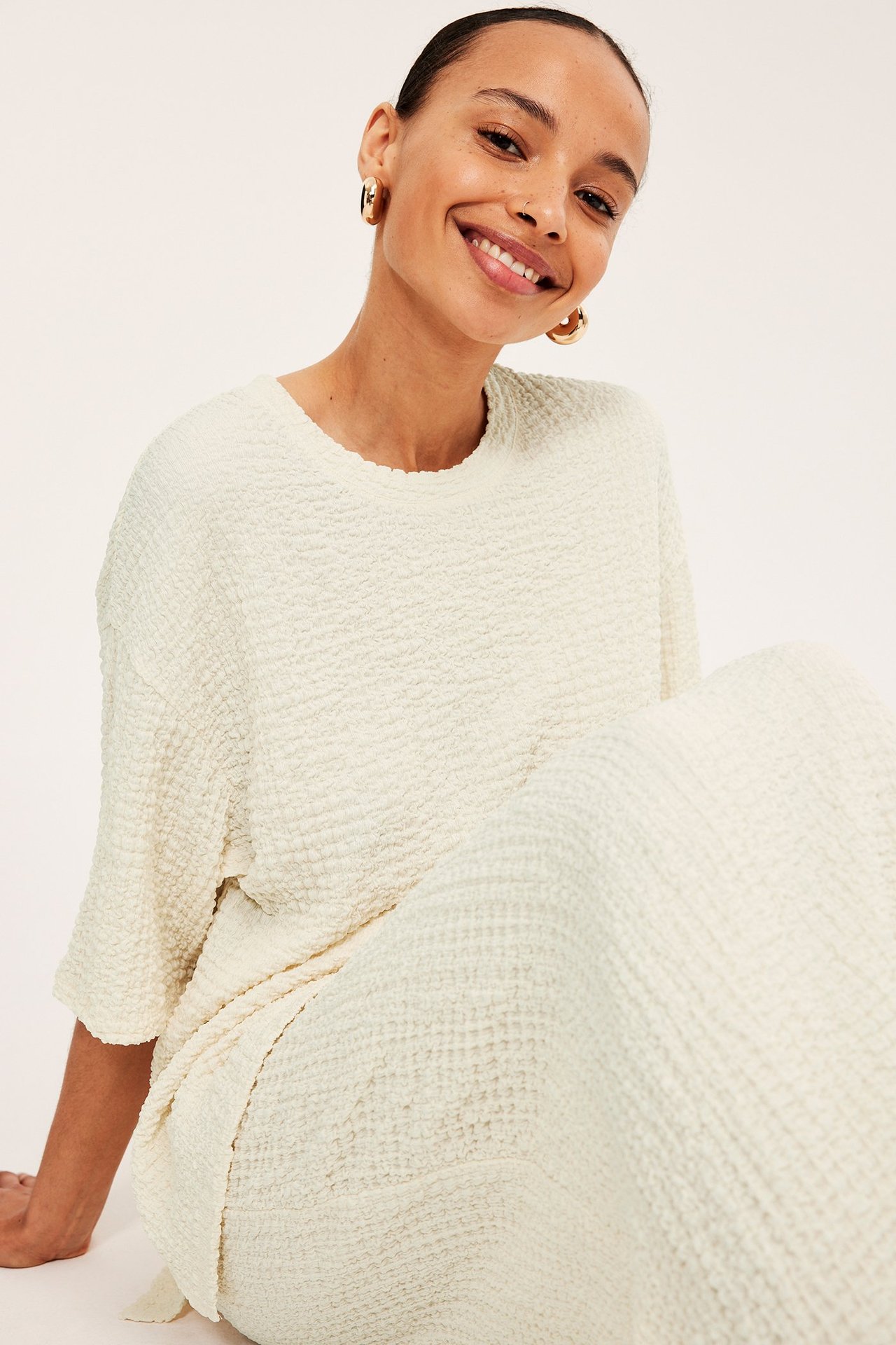 Uśmiechnięta kobieta w kremowym topie o strukturze, pasujących spodniach i złotych kolczykach.