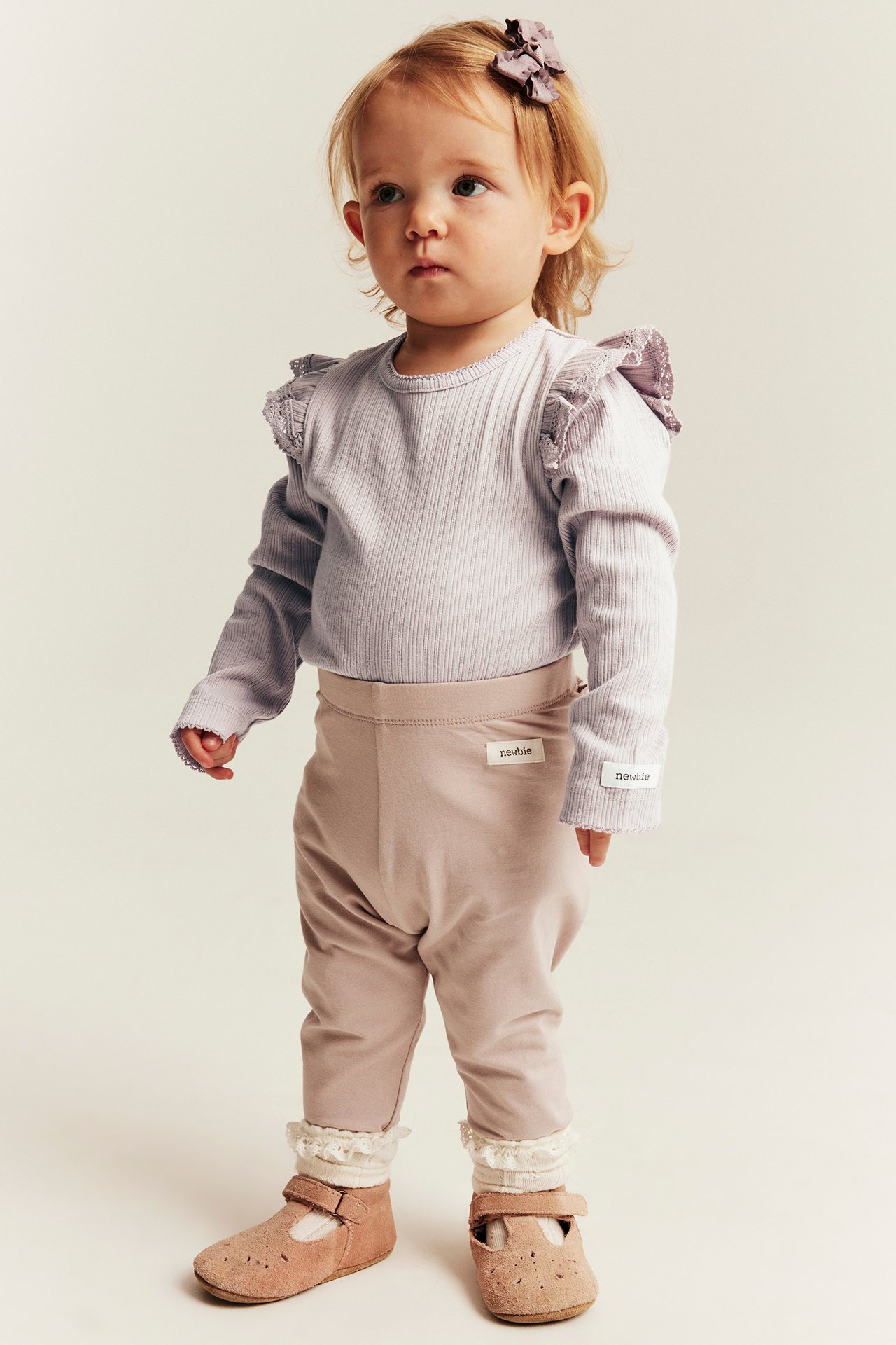 Baby i lilla ribbet volangtopp, brune leggings, hvite strømper, brune sko, lilla sløyfe.
