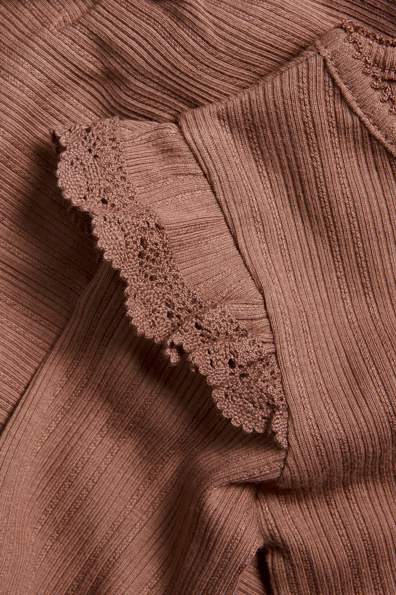 Nærbilde av en mørk rosa ribbet babybody med volanger og blondekant på skulderen.