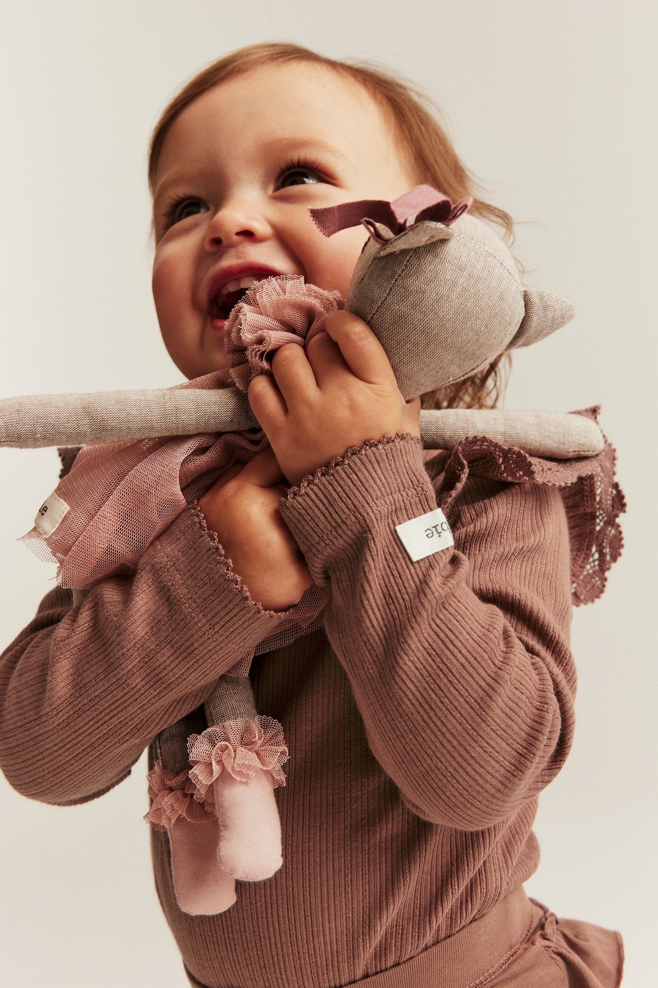 Lähikuva vauvasta tummanroosassa ribbipaidassa pitsiröyhelöillä, pitäen kangasnukkea.