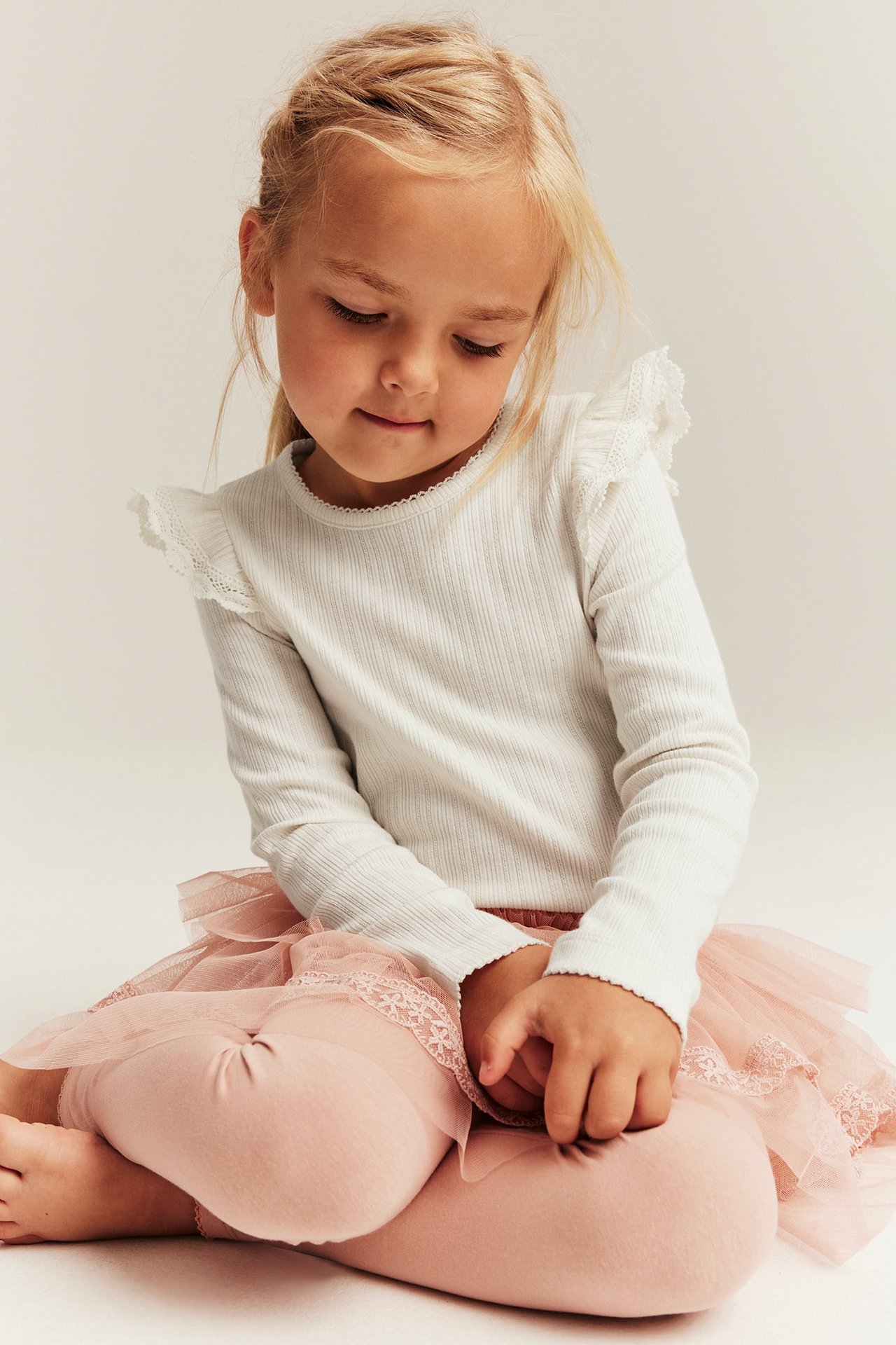 Widok z przodu: dziecko w kremowym prążkowanym topie z falbankami i koronką, oraz różowych tiulowych legginsach.