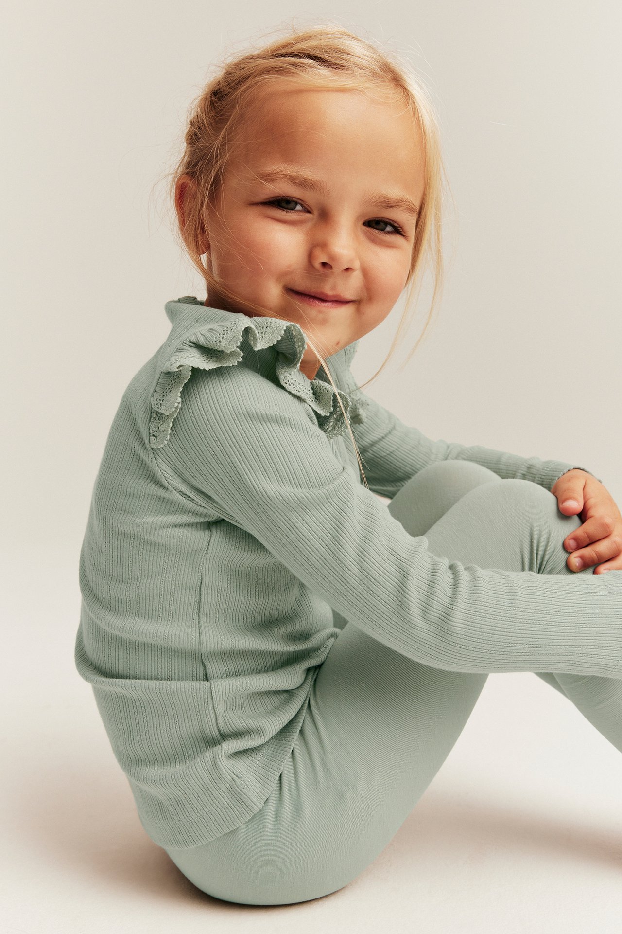 Frontbilde av et barn i lysegrønn ribbet topp med volanger og blondekanter på skuldrene, og leggings.
