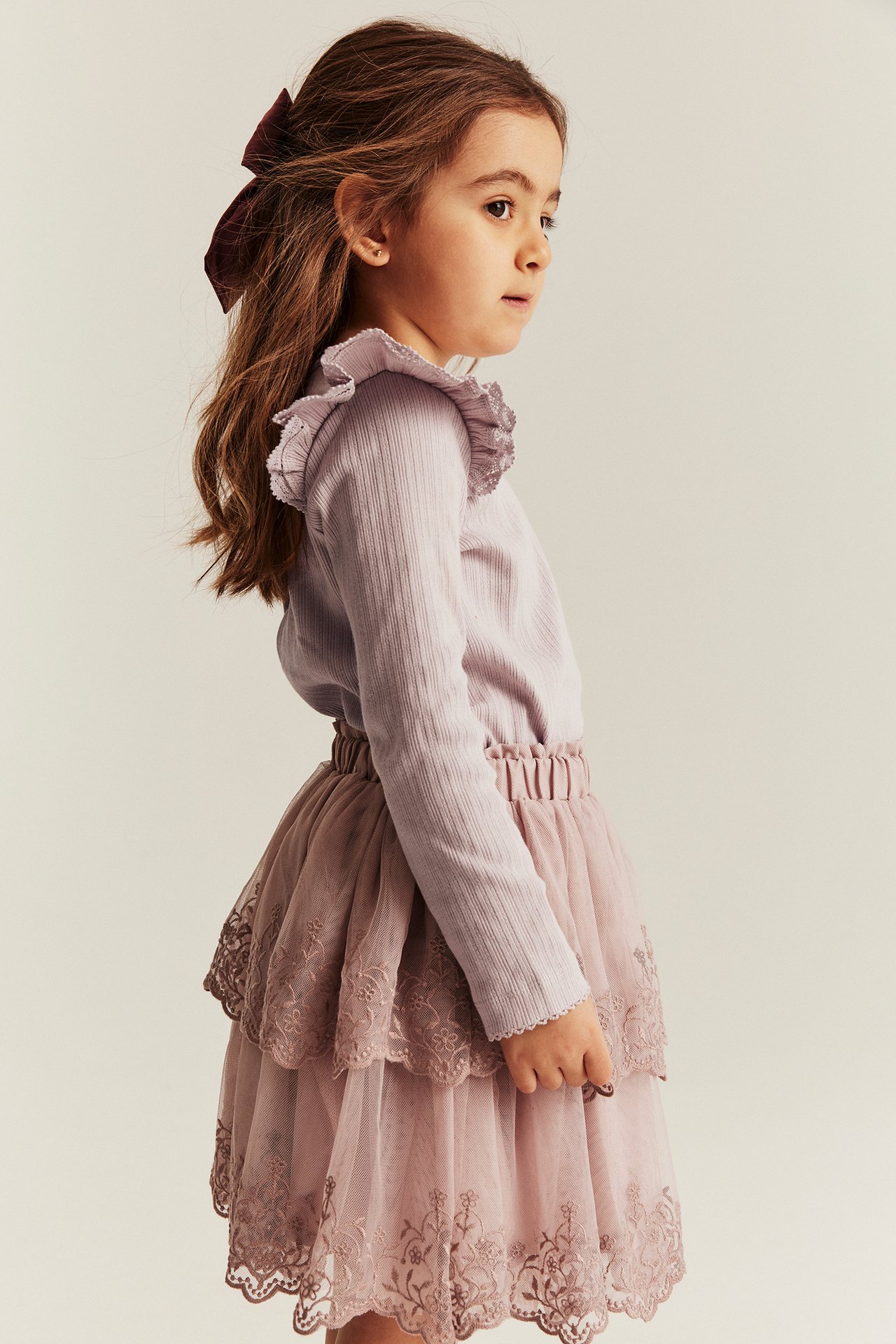 Sidevisning av barn i lyslilla ribbet topp med volanger, rosa skjørt og mørkerød sløyfe.