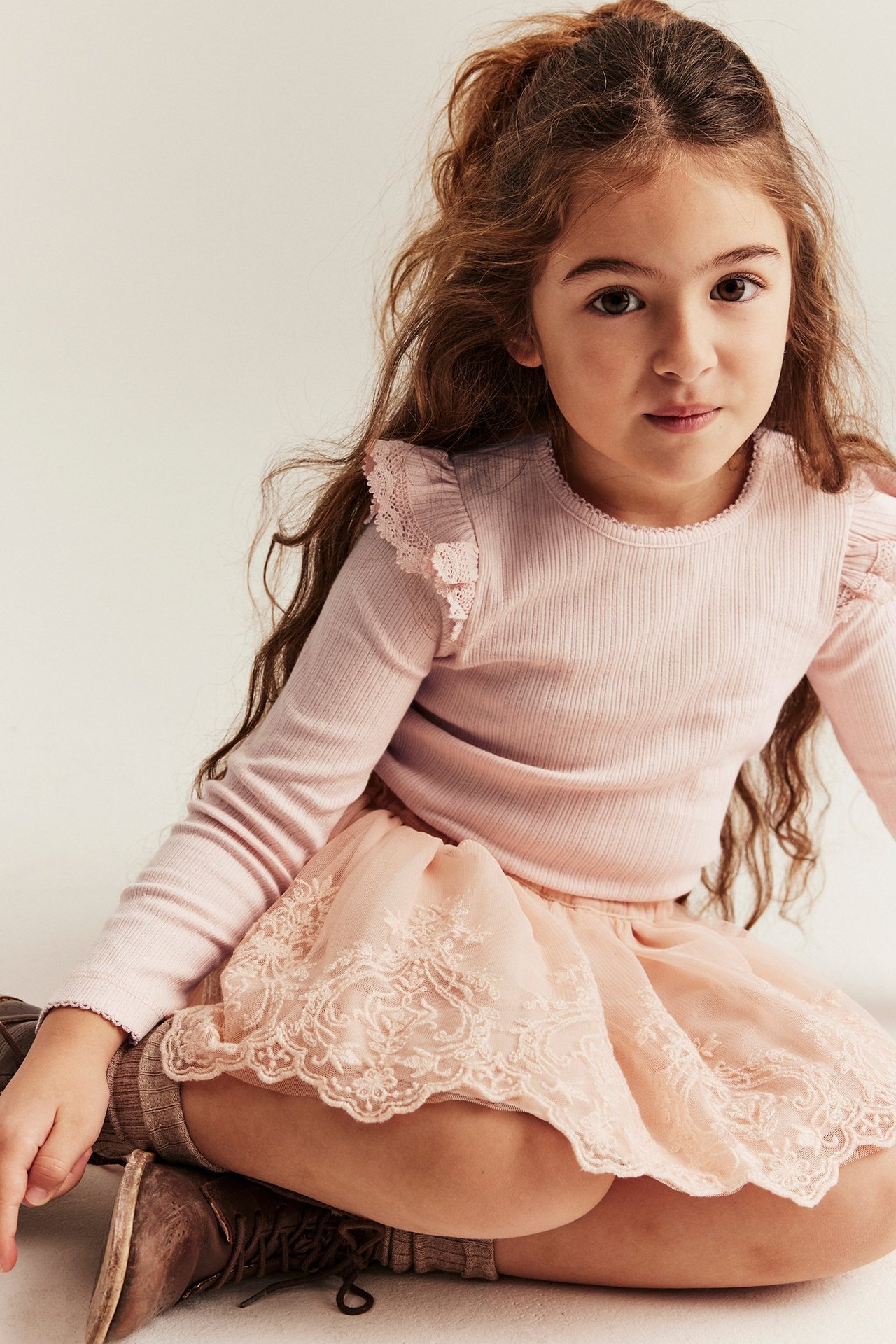 Edestä: Pikkutyttö vaaleanpunaisessa ribbipaidassa pitsiröyhelöin, brodeeratussa hameessa ja ruskeissa kengissä.