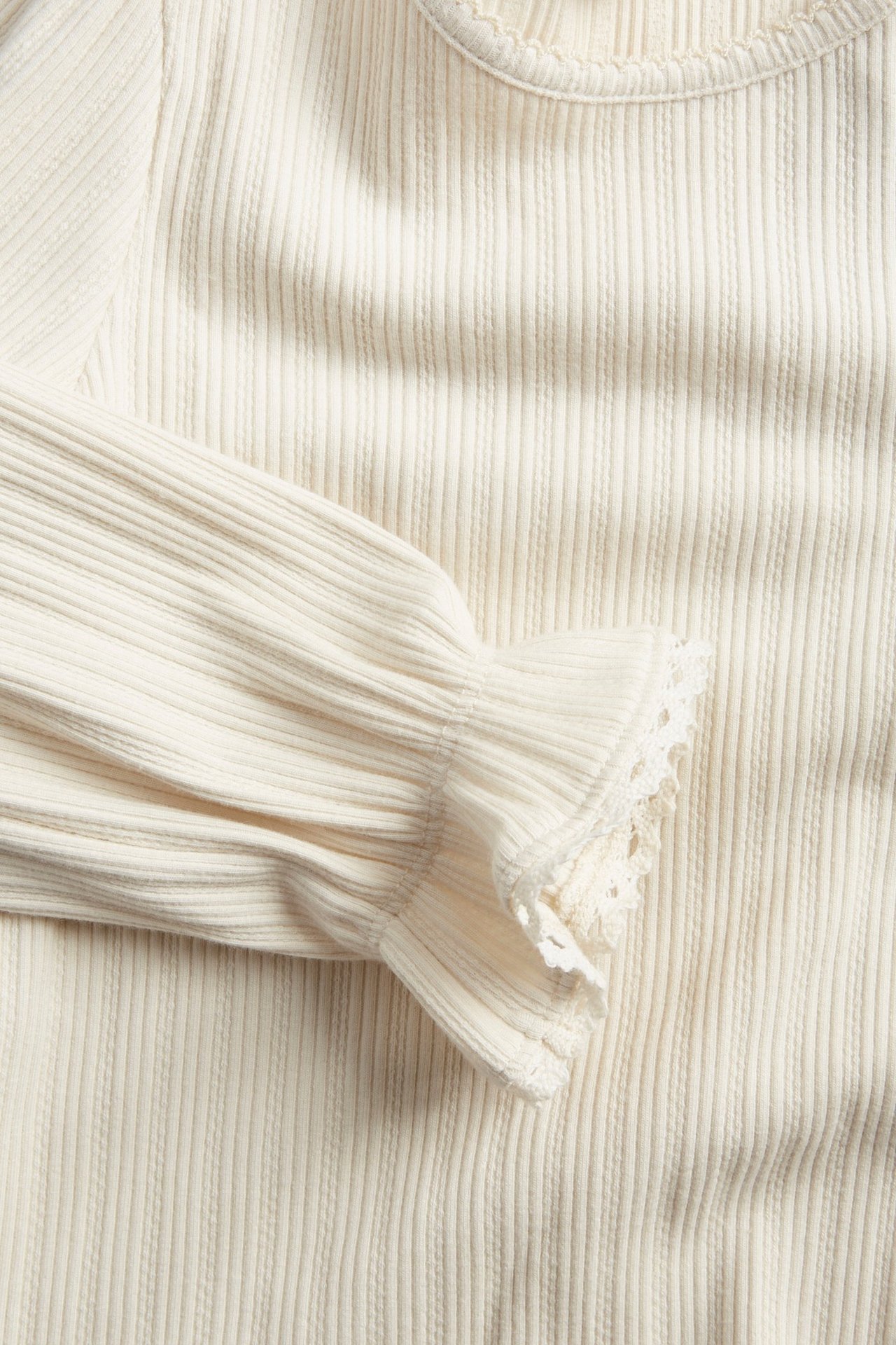 Zbliżenie jasnobeżowego prążkowanego topu z falbankami, koronkowymi mankietami i dekoltem z pikotką.