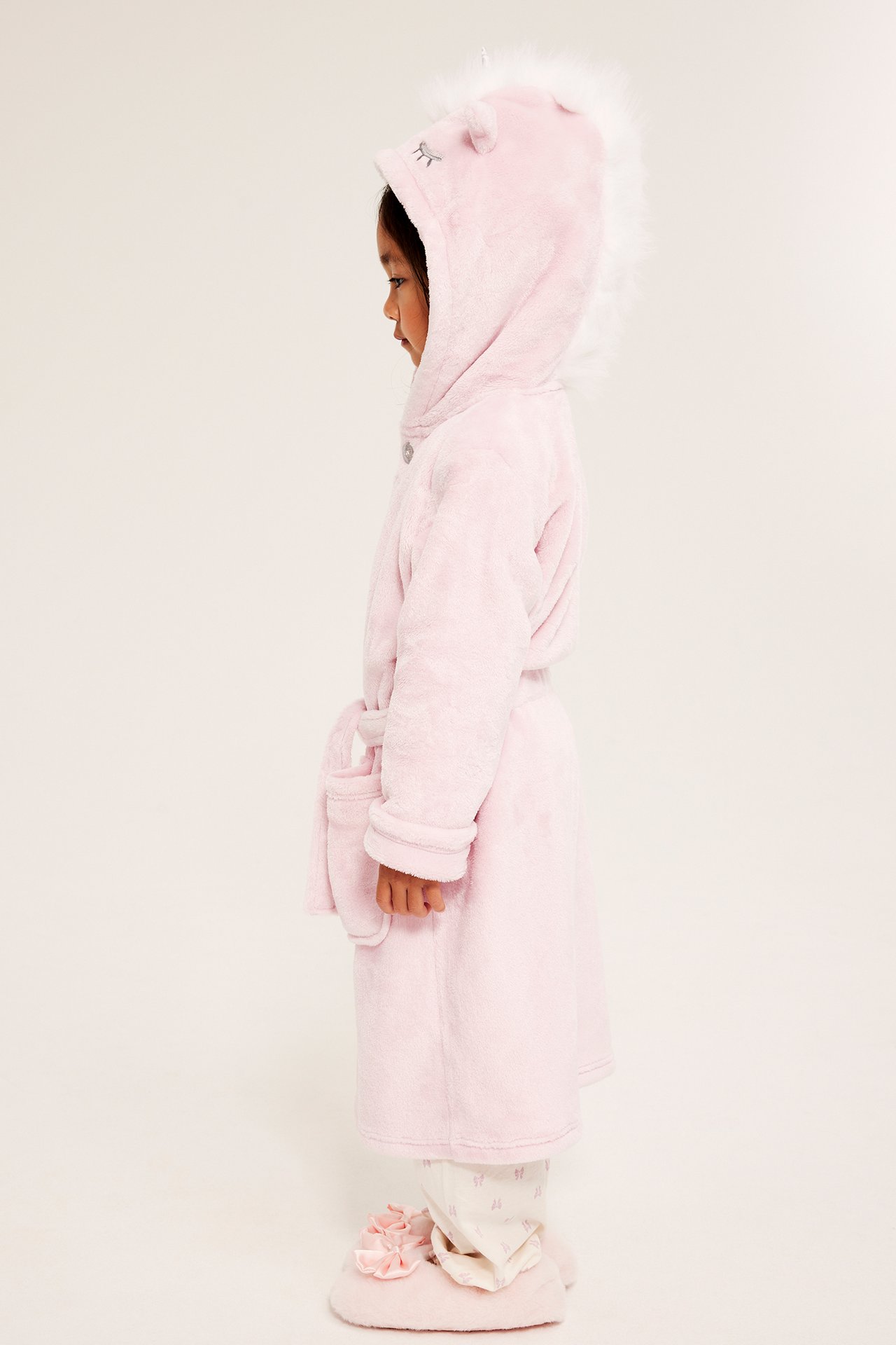 Sivukuva lapsesta yllään vaaleanpunainen fleece-yksisarvis-aamutakki, kuviolliset pyjamat ja vaaleanpunaiset tohvelit.