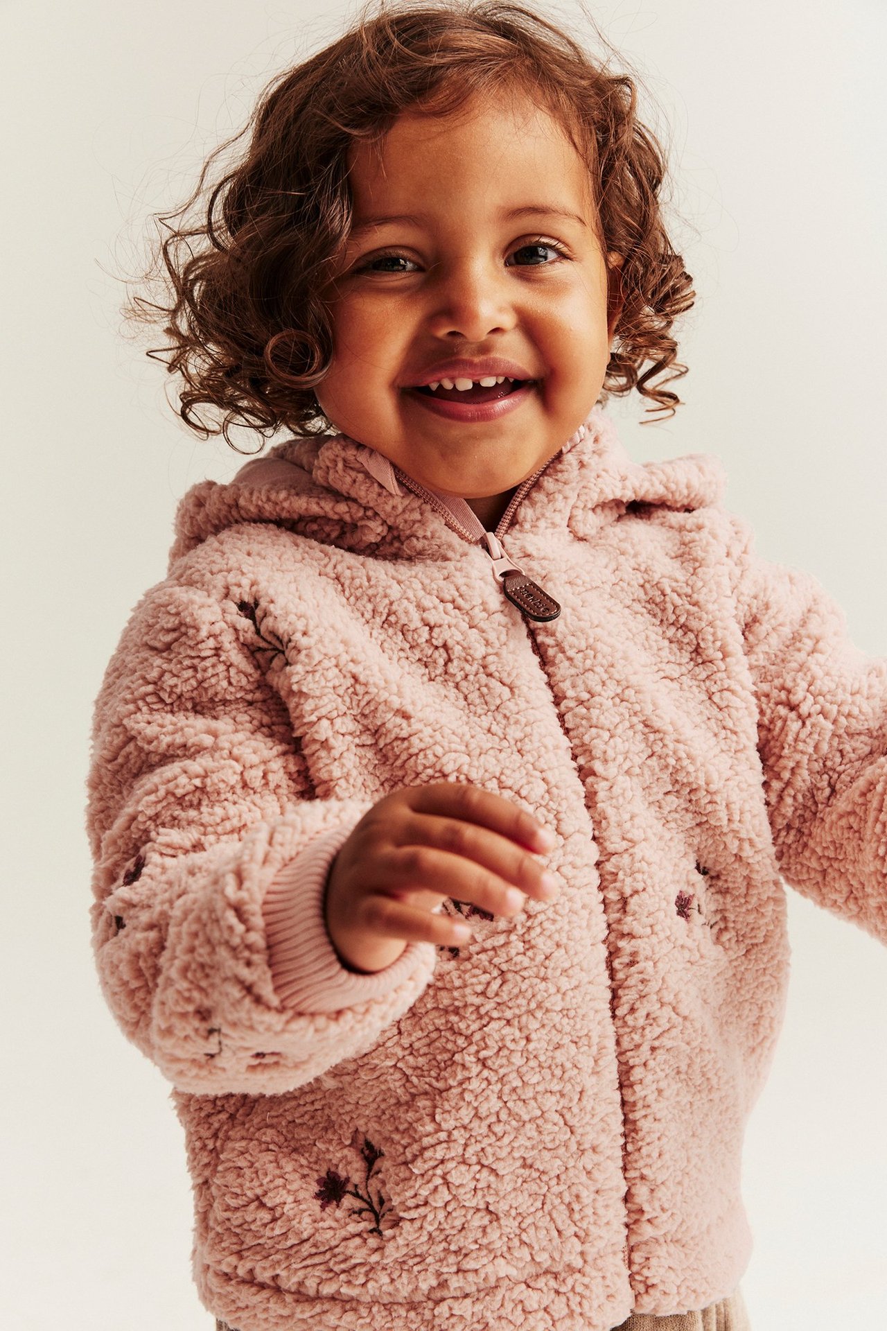 Frontvisning av et smilende barn i en rosa pilejakke med blomsterbroderi og glidelås.