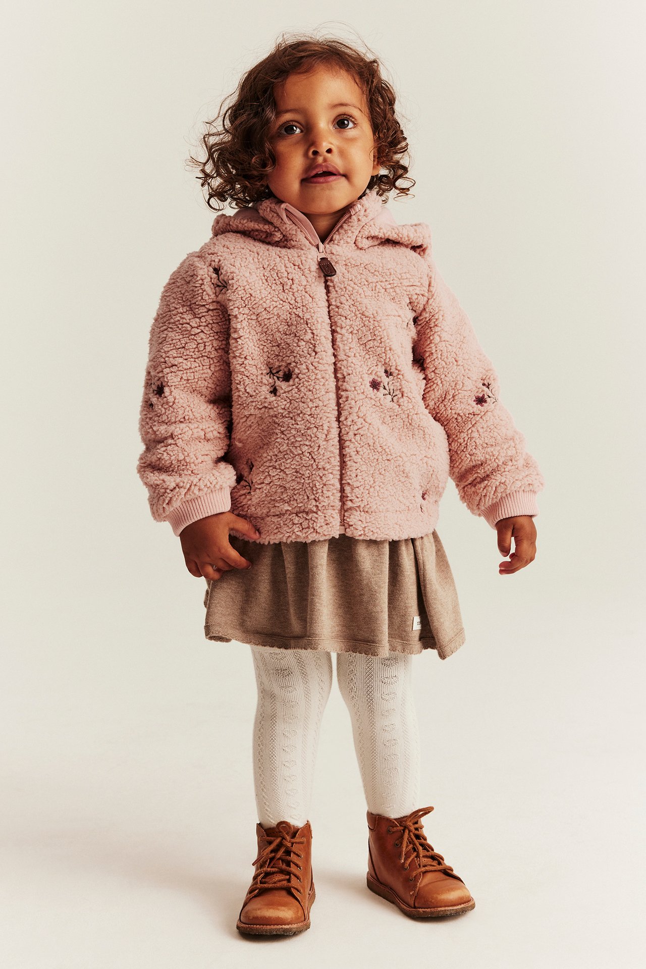 Framifrån: Barn i rosa teddyjacka med huva och blommor, beige kjol, vita strumpbyxor, bruna kängor.