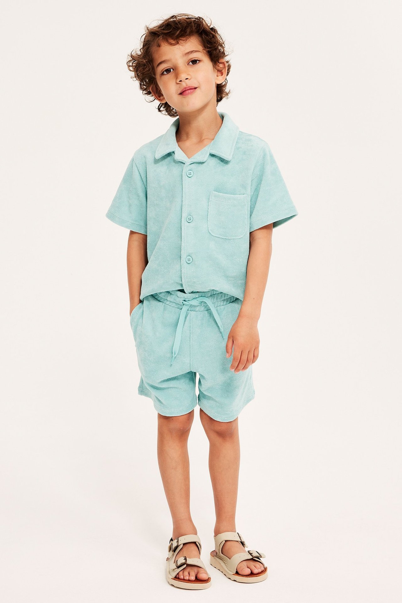Frontvy av ett barn i ljusgröna frottéshorts med dragsko, matchande skjorta och beiga sandaler.