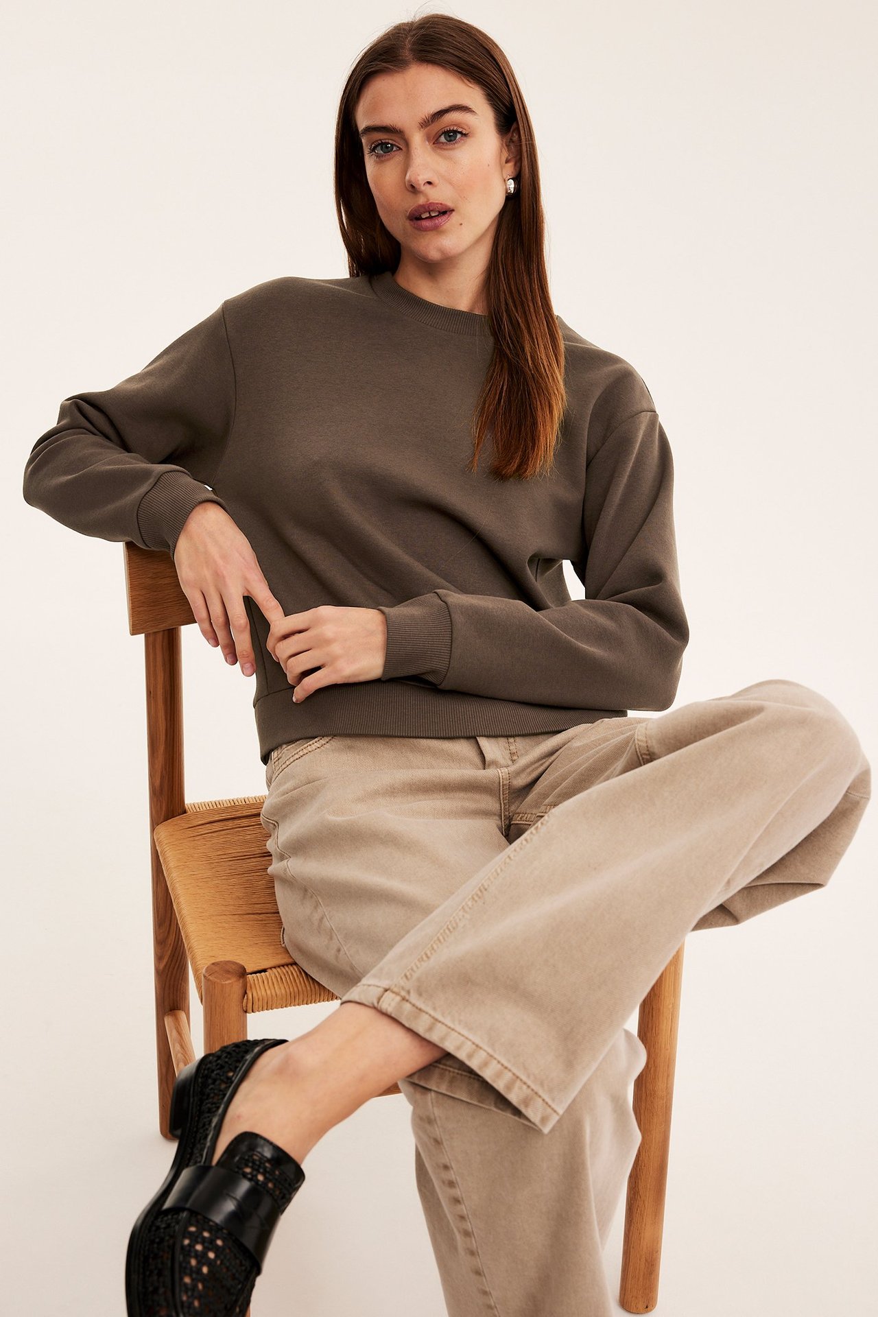 Widok z przodu: kobieta w ciemnobrązowej bluzie, jasnobeżowych szerokich spodniach i czarnych plecionych mokasynach.