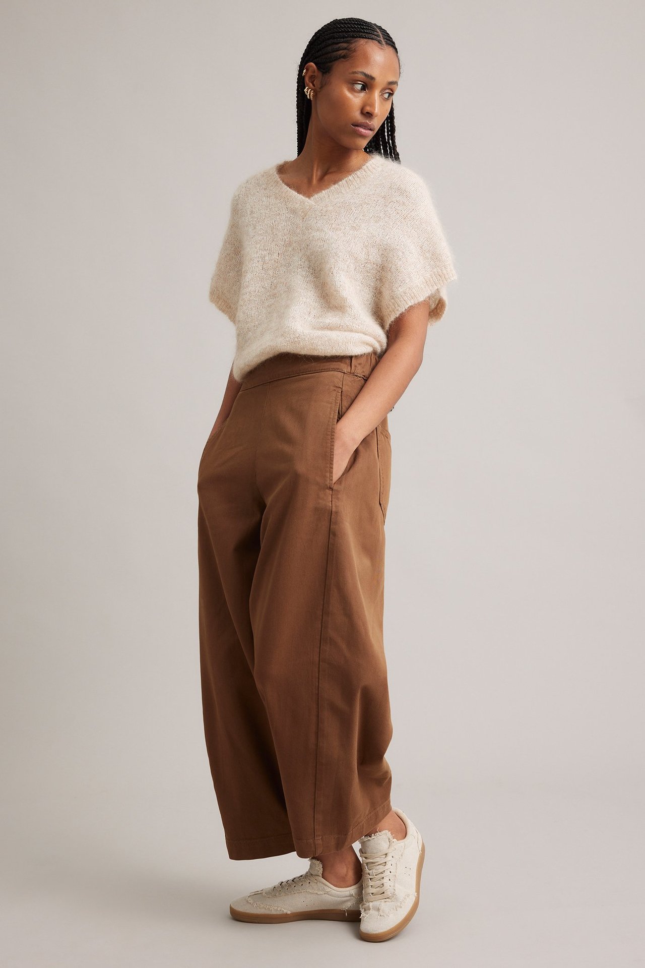 Sidovy av en kvinna i ljusbeige V-ringad stickad topp i mohairblandning, bruna vida byxor och ljusbeige sneakers.