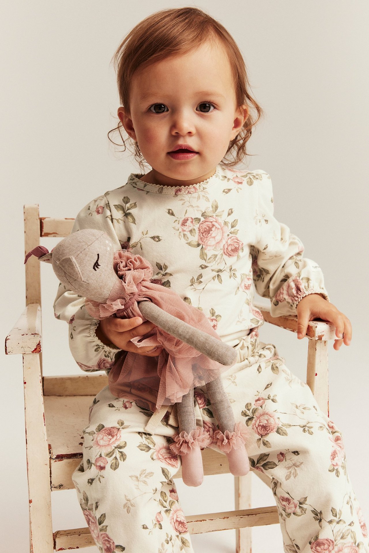 Widok z przodu: niemowlę w jasnobeżowym body w kwiaty z bufiastymi rękawami i pasujących spodniach, trzyma różową lalkę.