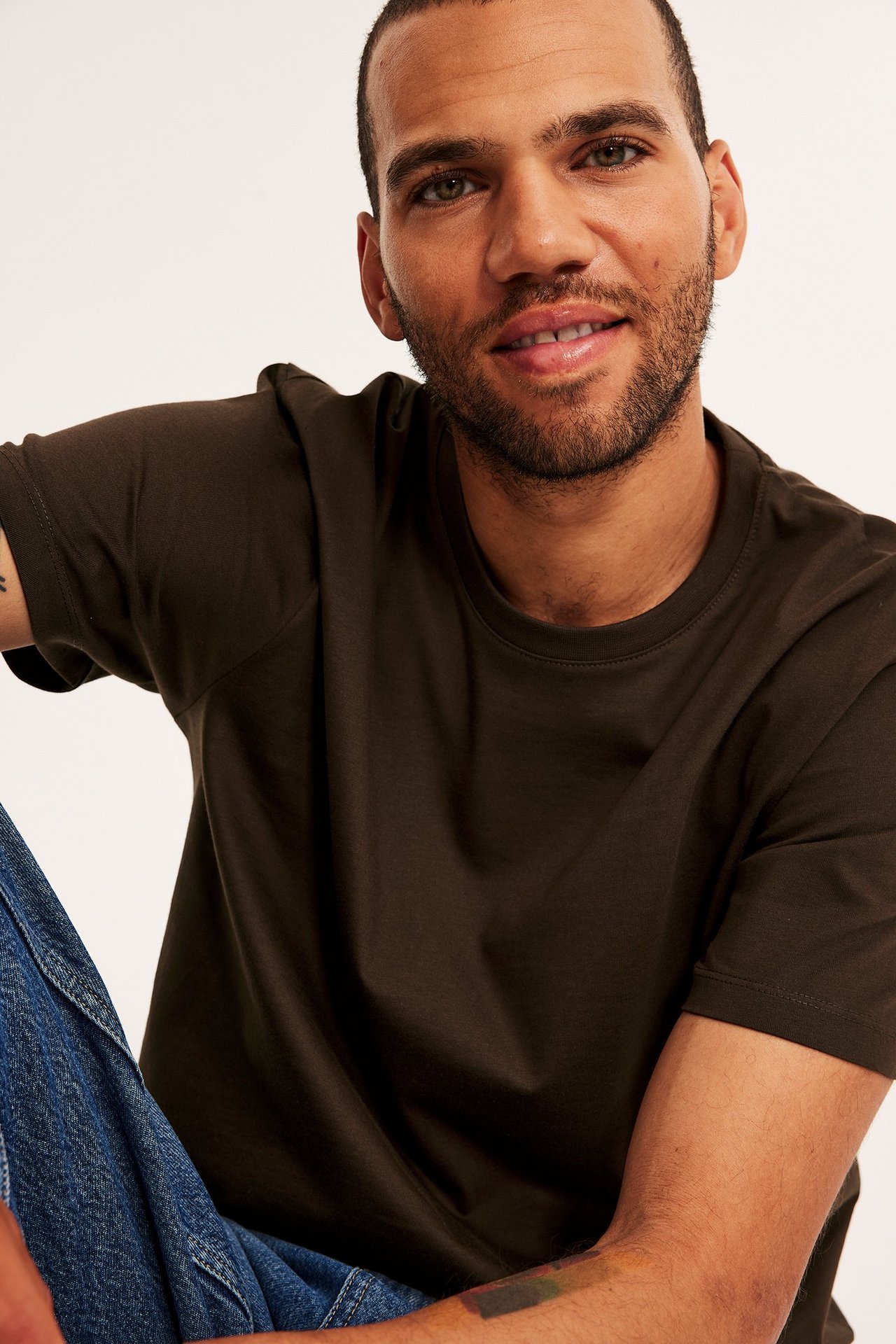 Lähikuva hymyilevästä miehestä, jolla on yksivärinen ruskea pyöreäkauluksinen t-paita ja siniset farkut.