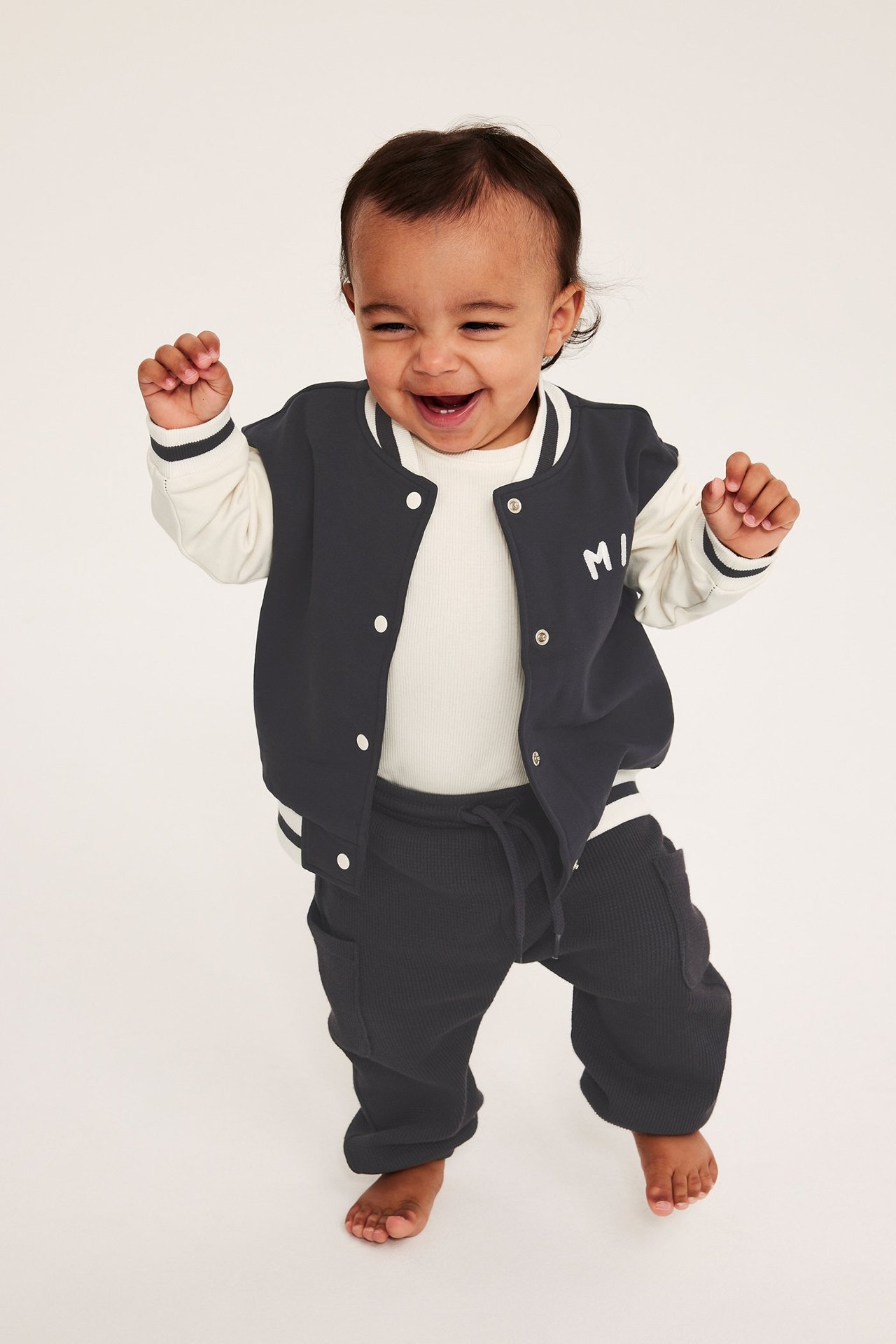 Leende baby i mörkgrå baseballjacka med 'MINI' applikation, krämfärgad ribbad topp och mörkgrå mjukisbyxor.