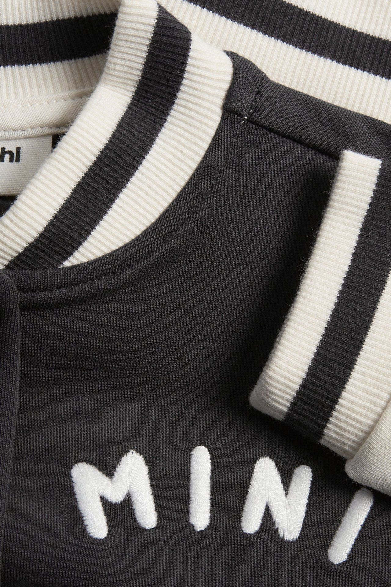 Zbliżenie czarnej kurtki bejsbolowej dla niemowląt z biało-paskowanym kołnierzem i mankietami, oraz białym haftem z przodu.