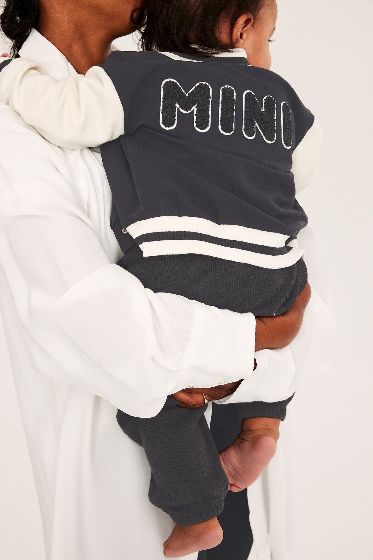 Bakifrån, baby i tvåfärgad baseballjacka med 'MINI'-applikation och mörkgrå ribbade byxor.
