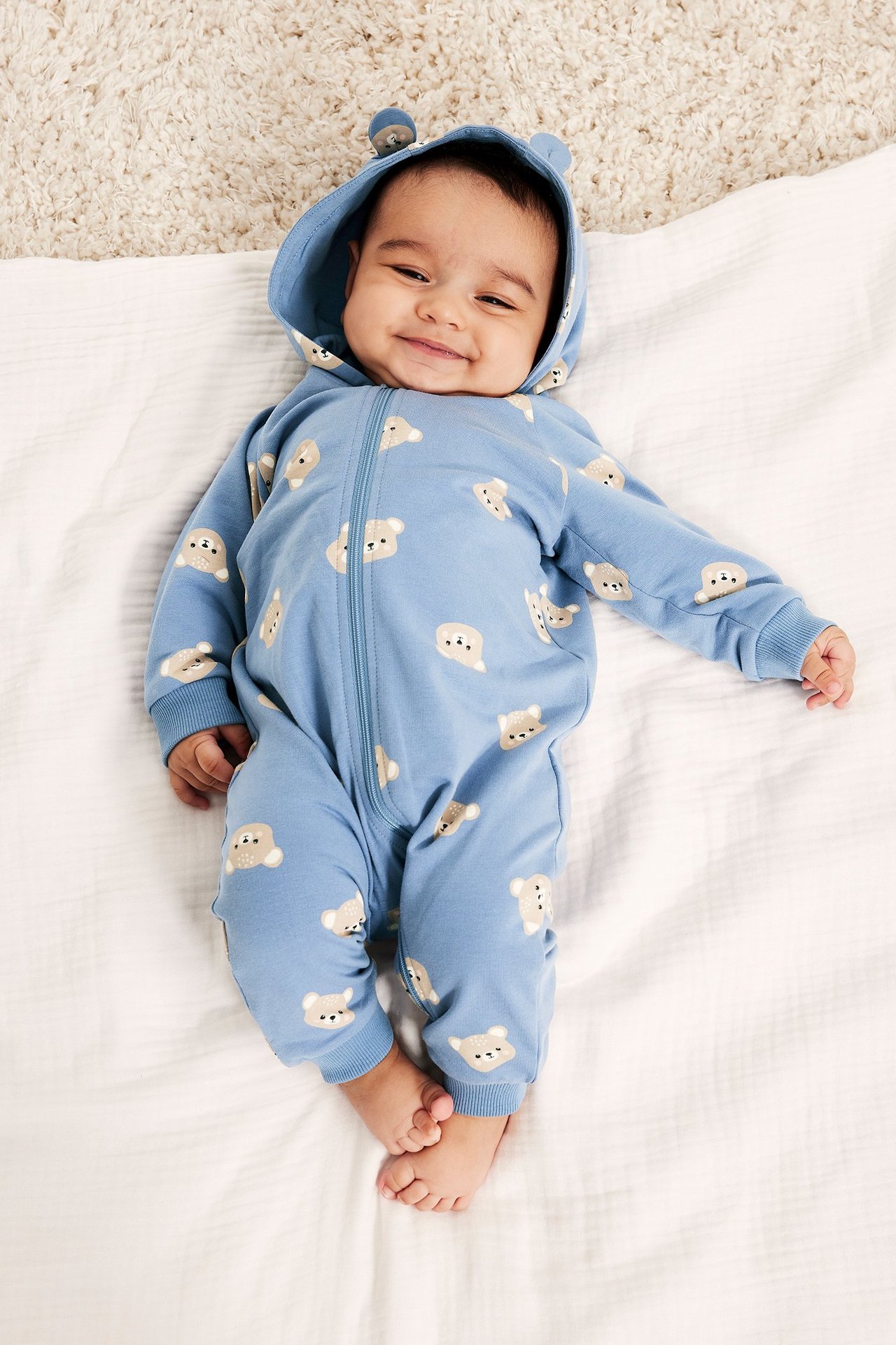 Uśmiechnięte dziecko w niebieskim kombinezonie z misiami, kapturem z uszami i zamkiem z przodu.
