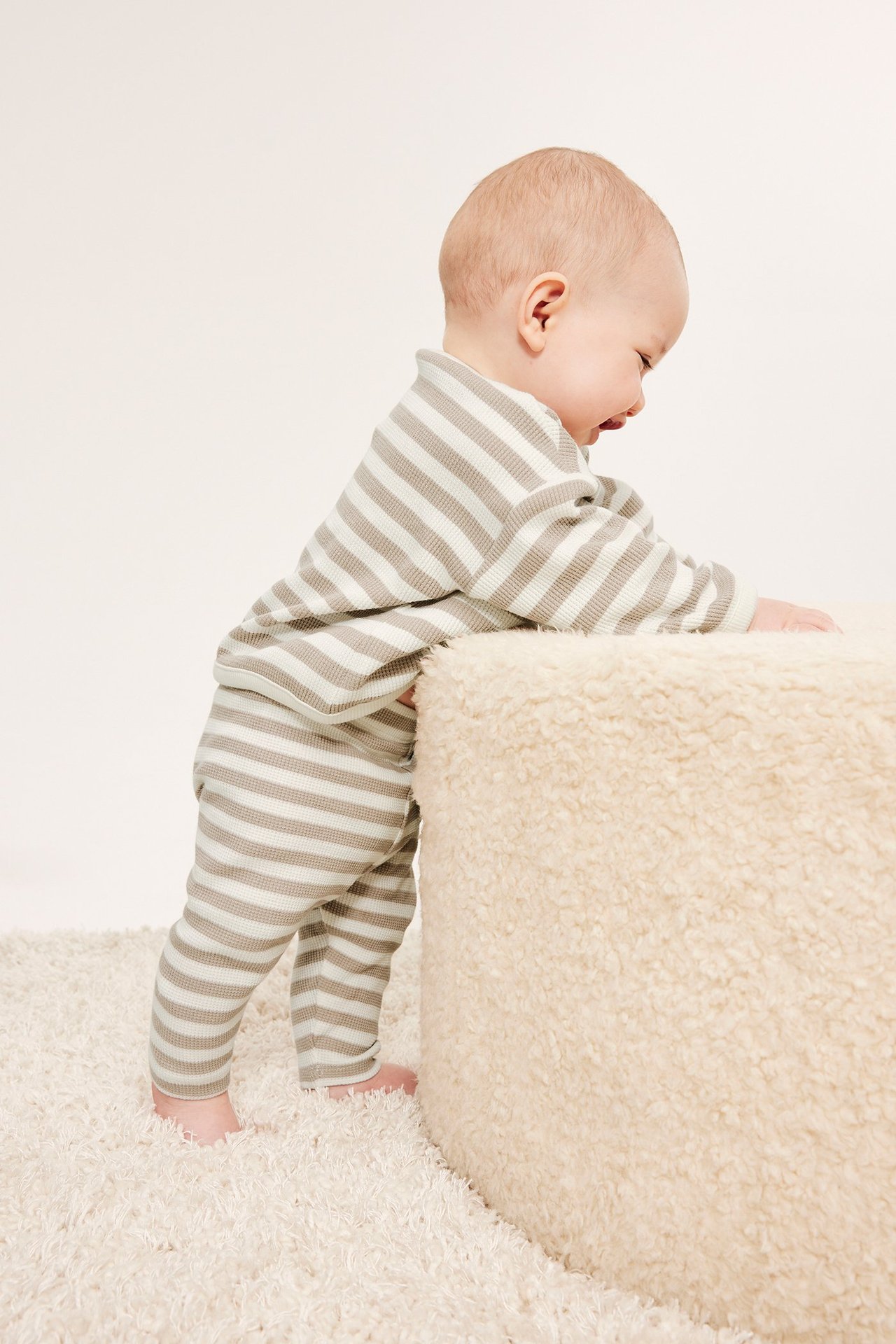 Sidebilde av baby i beige og hvitstripete vaffelstrikkede leggings og matchende langermet topp.
