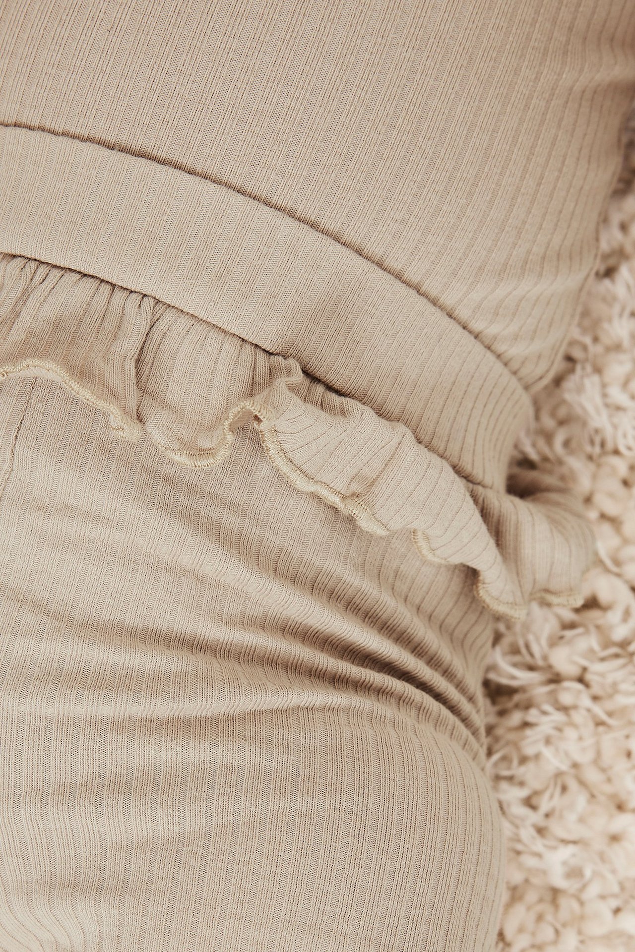 Nærbilde av beige ribbestrikkede baby leggings med volang i midjen, på et mykt teppe.