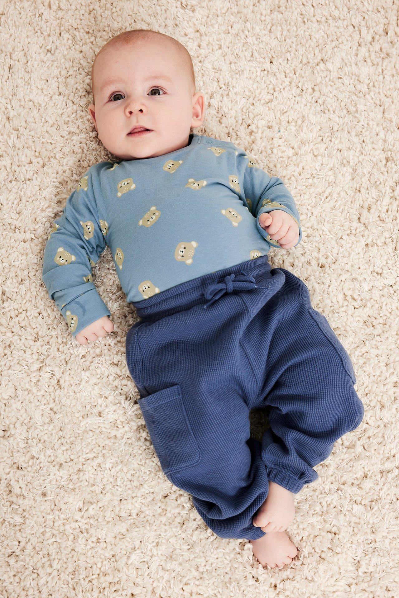 Ovenfra sett, en baby i lyseblå langermet body med bjørnemønster og mørkeblå vaffelstrikkede bukser.
