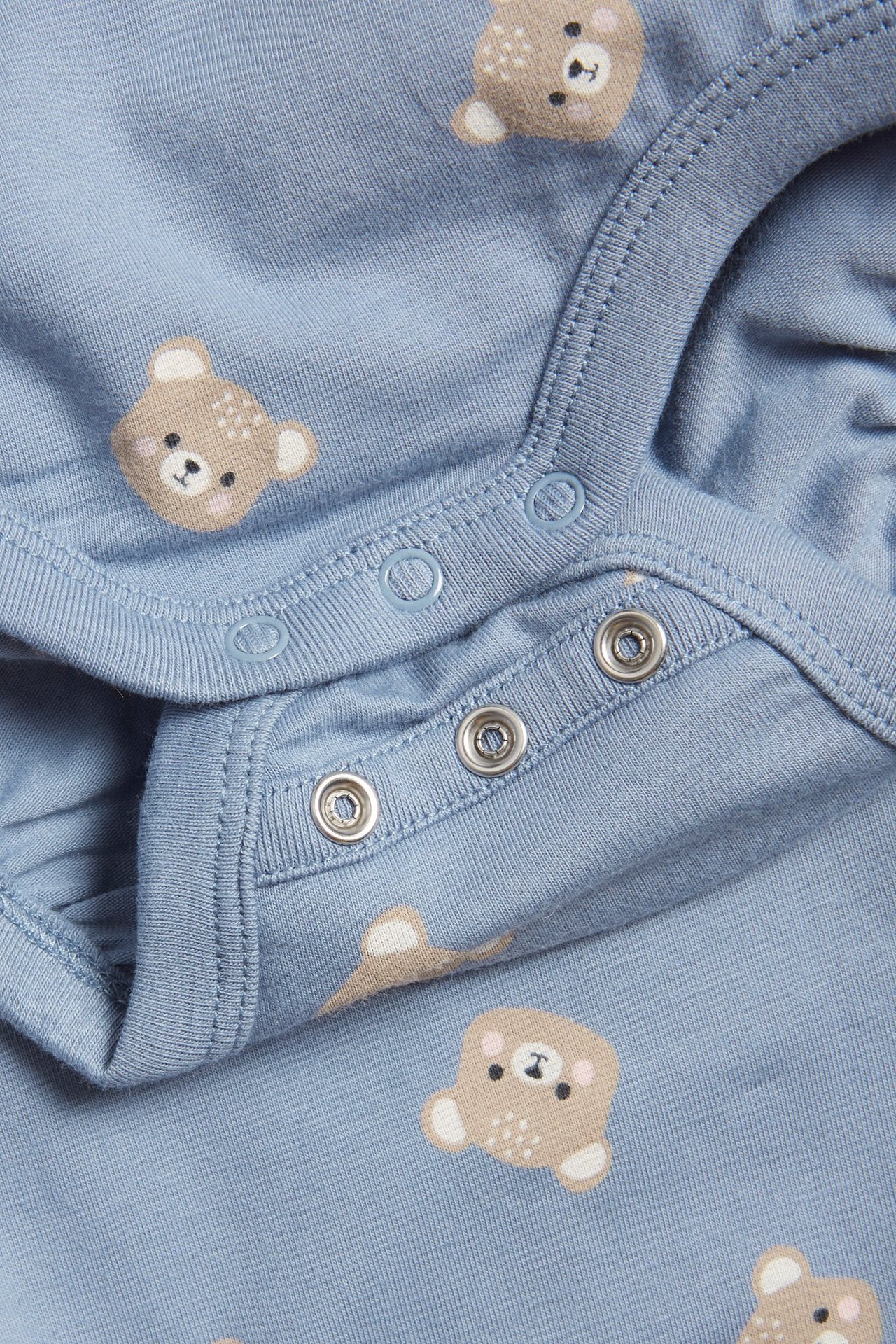 Zbliżenie niebieskiego bawełnianego body dla niemowląt z brązowym wzorem misia i zatrzaskami.