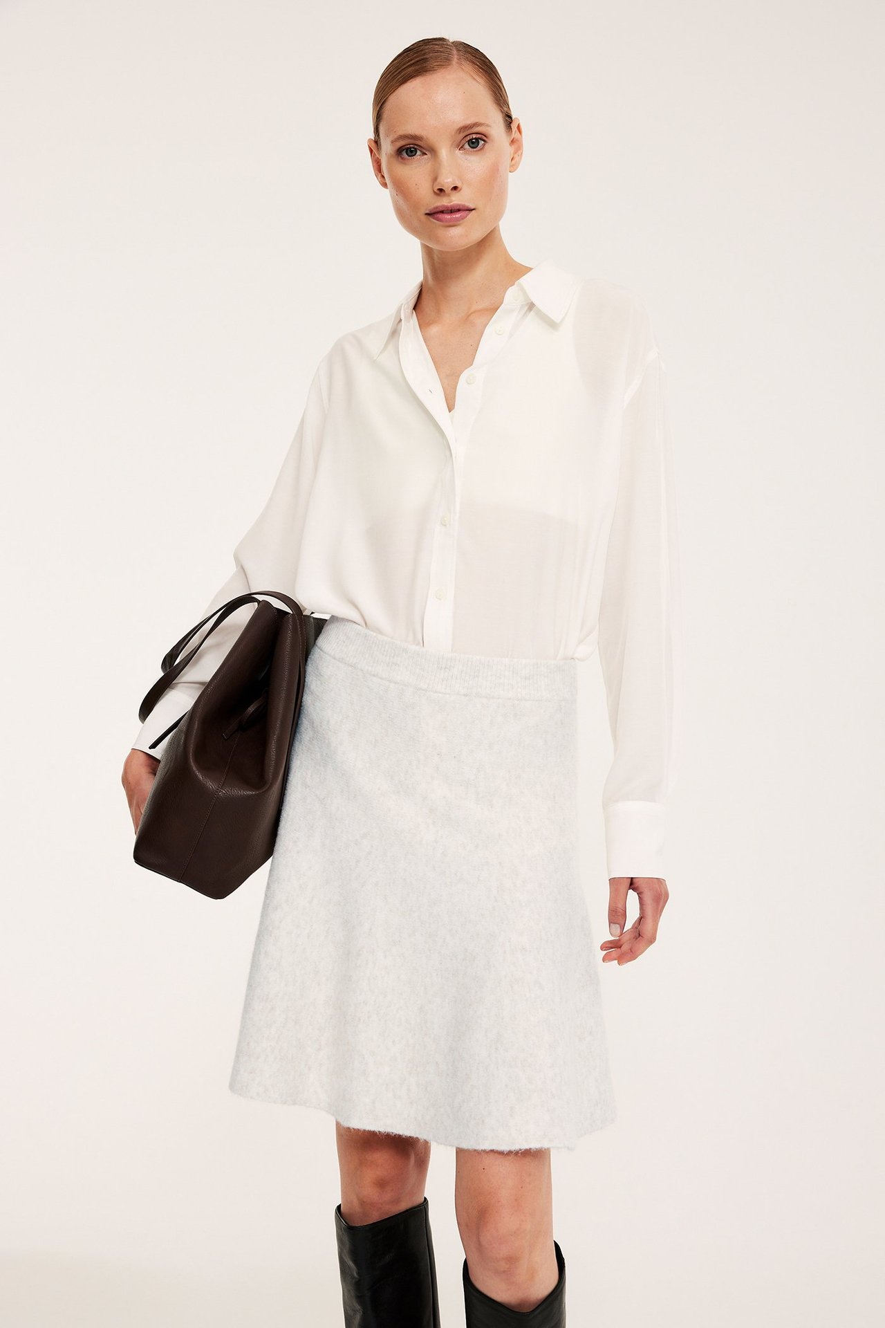 Edestä: Nainen yllään vaalea neulottu kellohame, valkoinen paita, pitää ruskeaa käsilaukkua ja mustia saappaita.