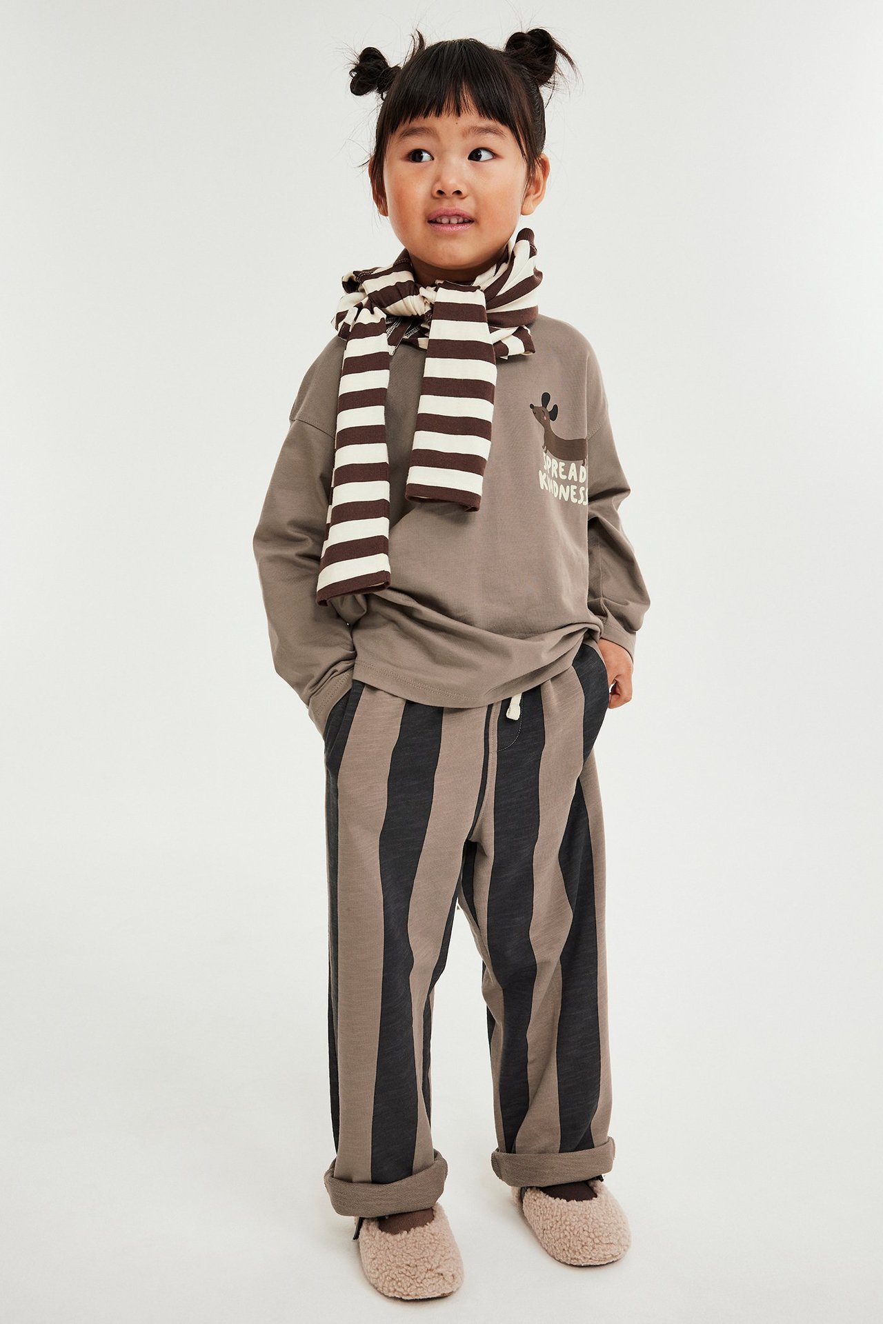 Front: Barn i brune blokkstripete joggebukser, taupe langermet topp, stripet skjerf og fluffy tøfler.