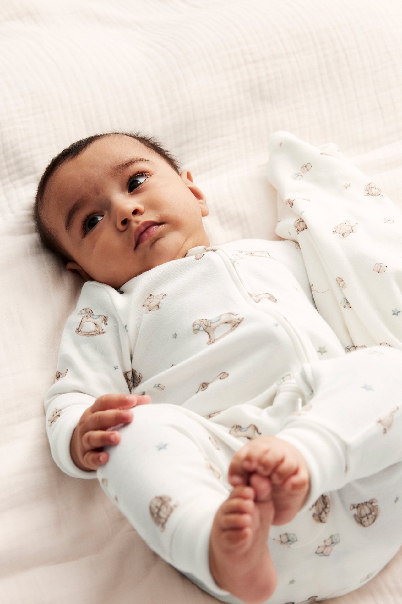 Edestä, vauva yllään luonnonvalkoinen puuvillapyjama lelukuvioilla ja kaksisuuntaisella vetoketjulla.