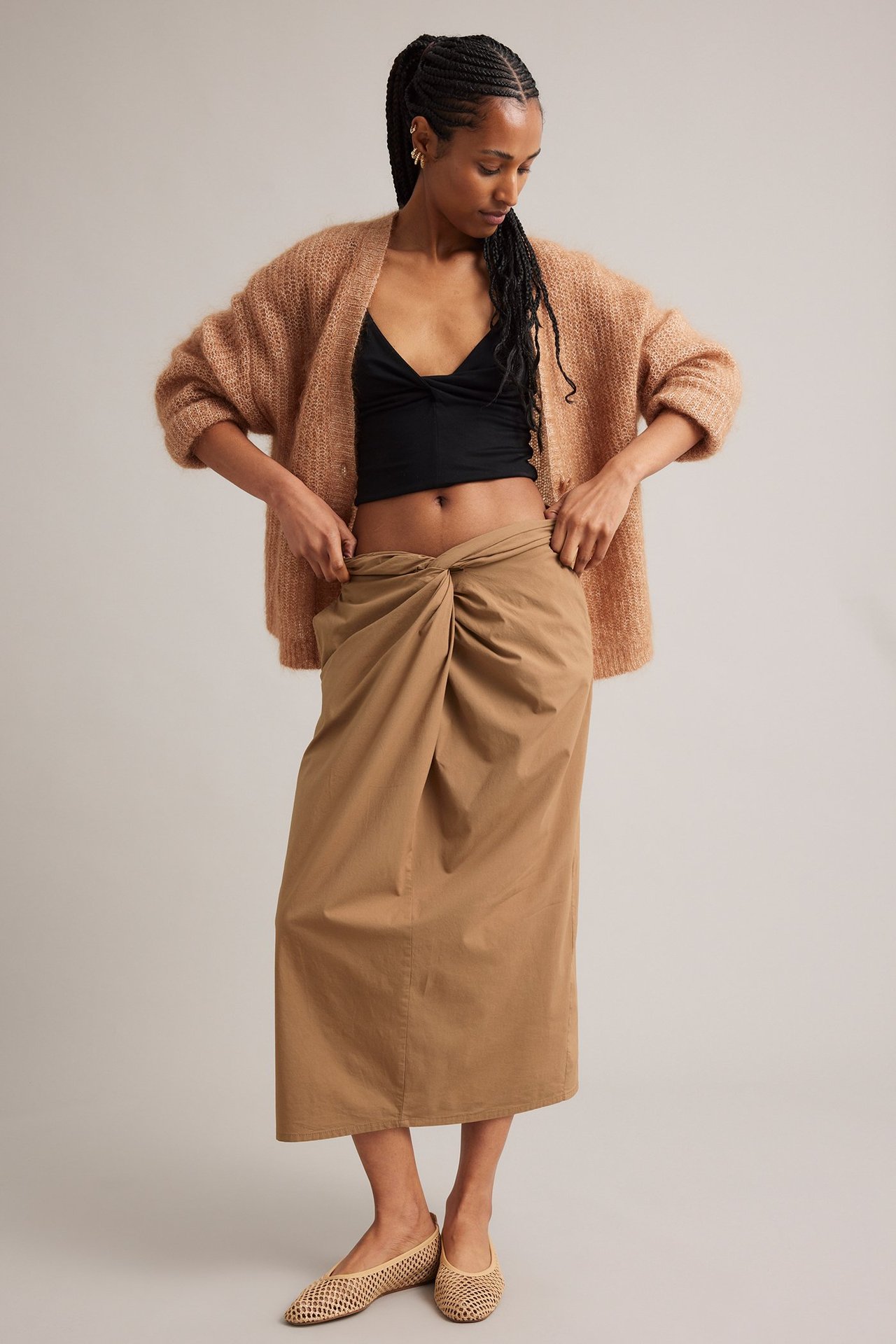 Frontalvisning: Kvinne i lys brunt midiskjørt med knute foran, svart crop top og lys brun cardigan.
