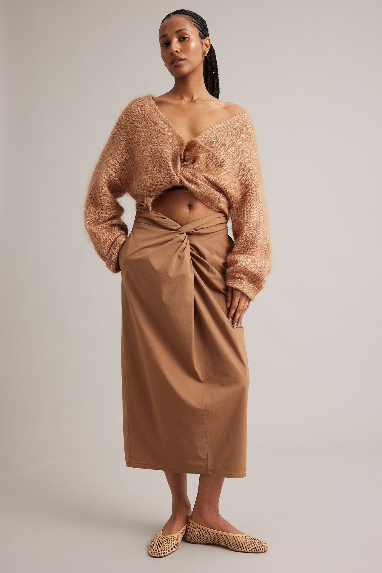 Frontvisning: Kvinne i brunt midiskjørt med knutedetalj, lys brun strikkegenser og beige vevde flate sko.