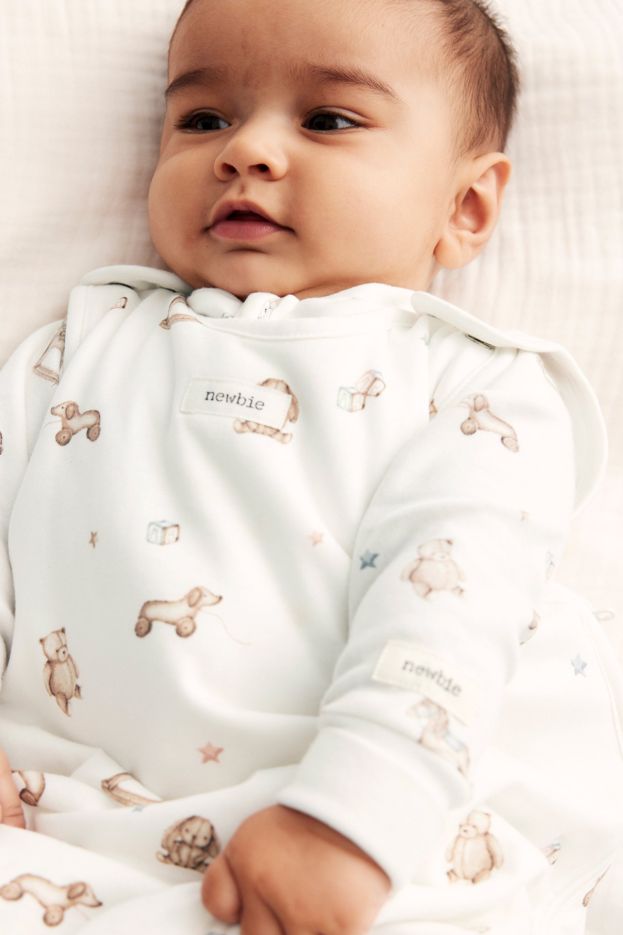 Närbild på en baby i en offwhite sovpåse med leksaksmönster och tryckknappar vid axlarna.