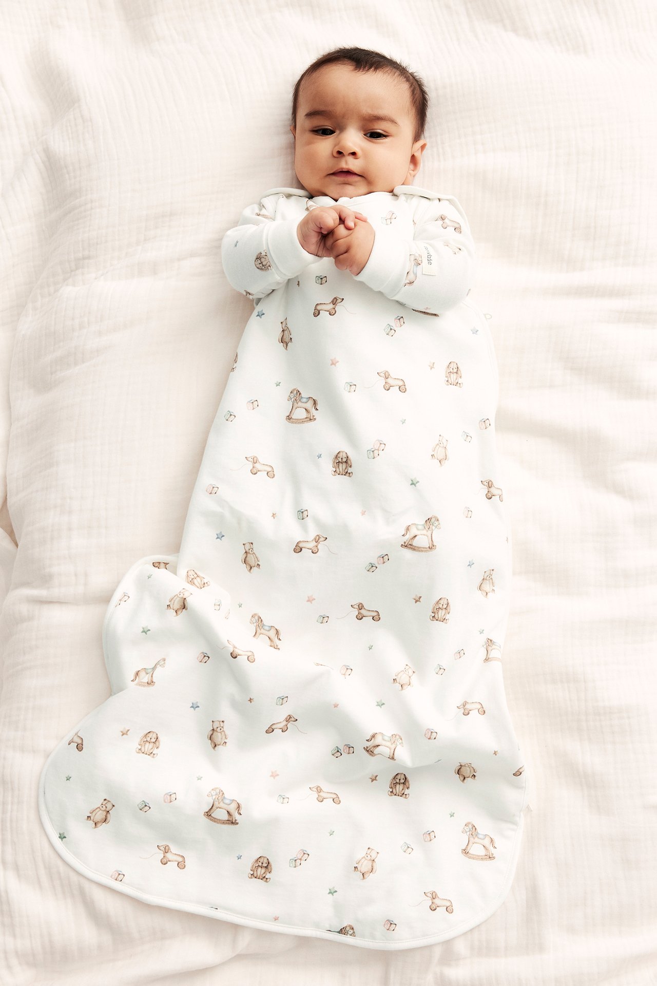 Uppifrån vy av en baby i en offwhite långärmad sovpåse med gulligt leksaksmönster och tryckknappar.