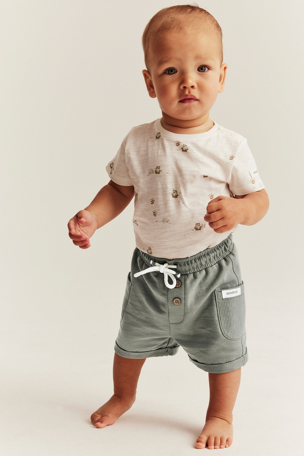 Edestäpäin, vauva yllään vihreät slub-trikooshortsit kiristysnyörillä ja luonnonvalkoinen kuvioitu t-paita.