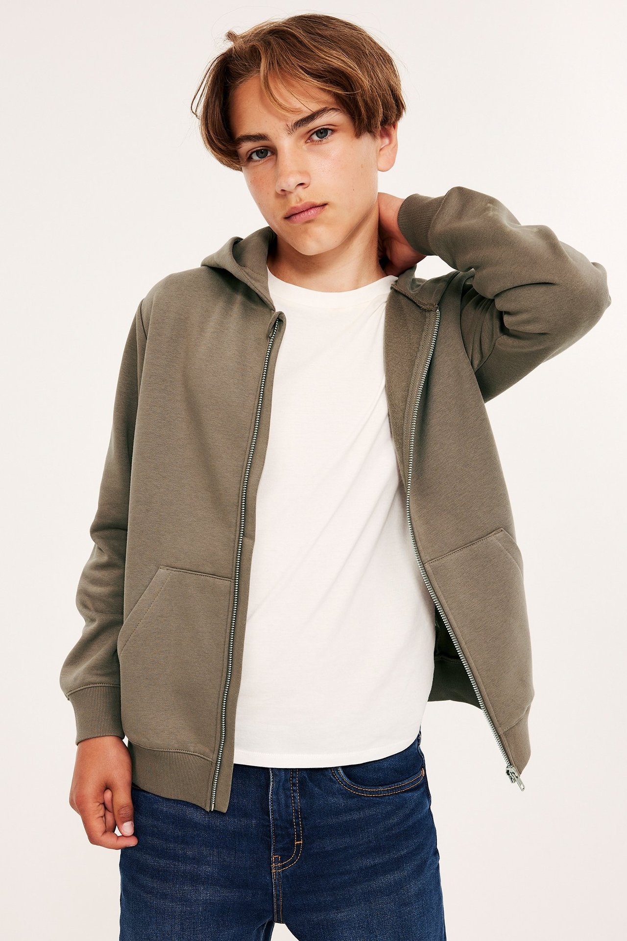 Framifrån: Kid bär en öppen brun hoodie med dragkedja över en vit t-shirt och blå jeans.