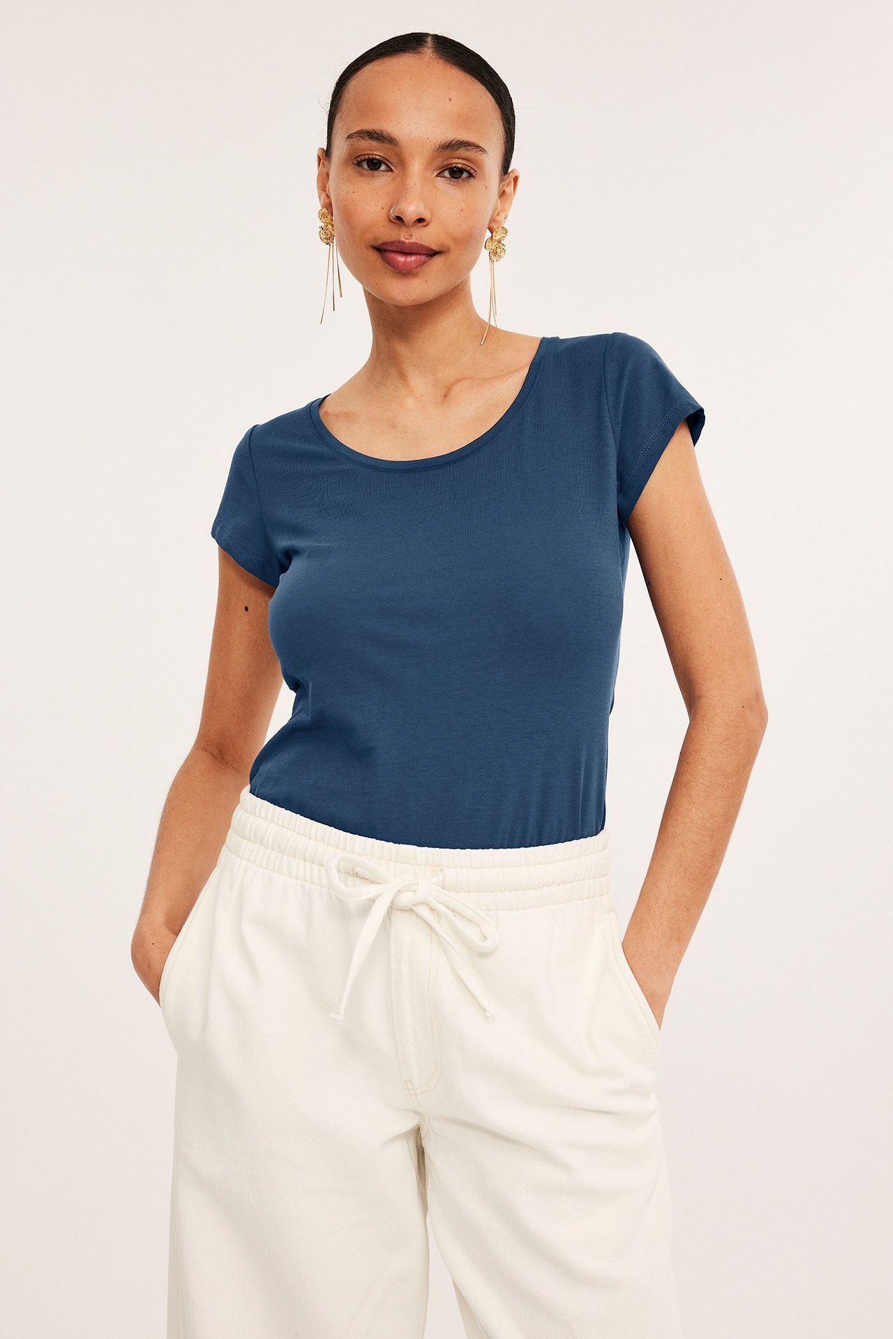 Frontbilde av en kvinne i en mørkeblå figurnær kortermet bomulls-t-skjorte, kremfargede bukser og gulløredobber.