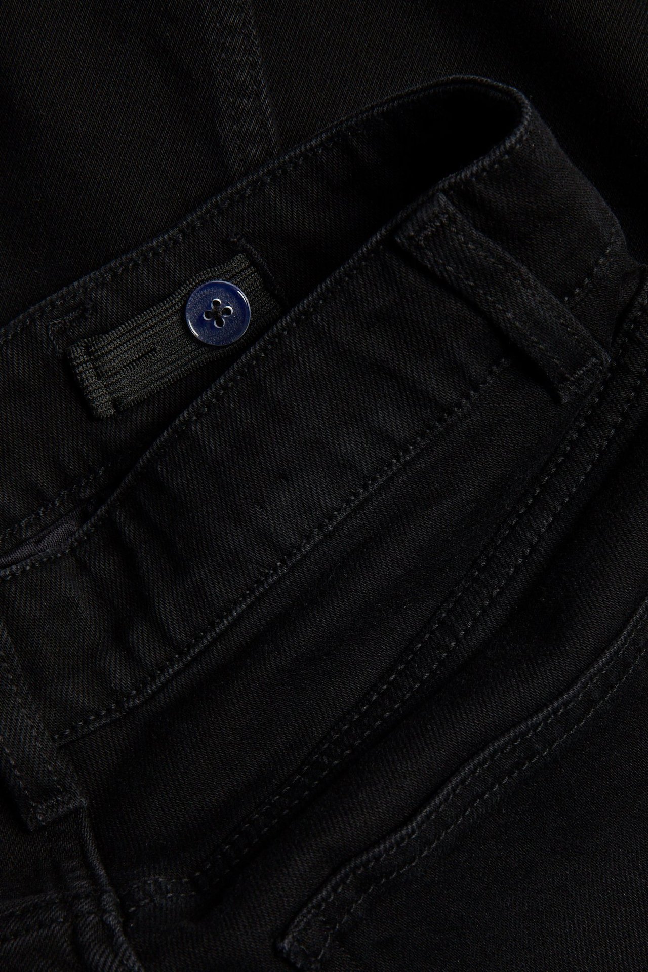 Närbild av svarta lösa jeans i denim med justerbar resår, medelhög midja och blå knapp.