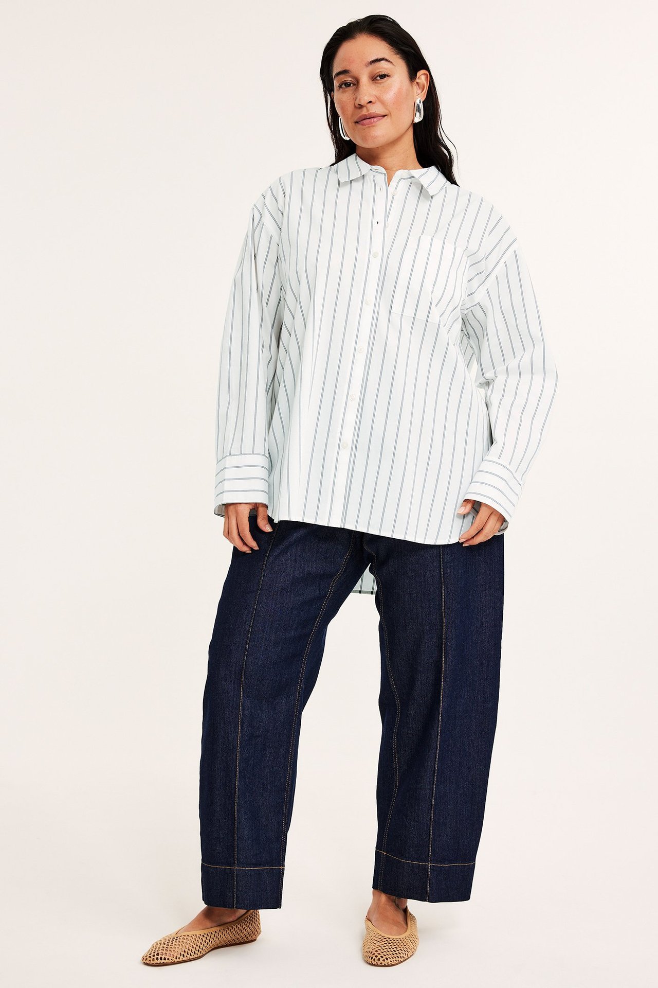 Frontvy av kvinna i offwhite randig oversized poplinskjorta, mörkblå vida byxor och beiga vävda ballerinaskor.