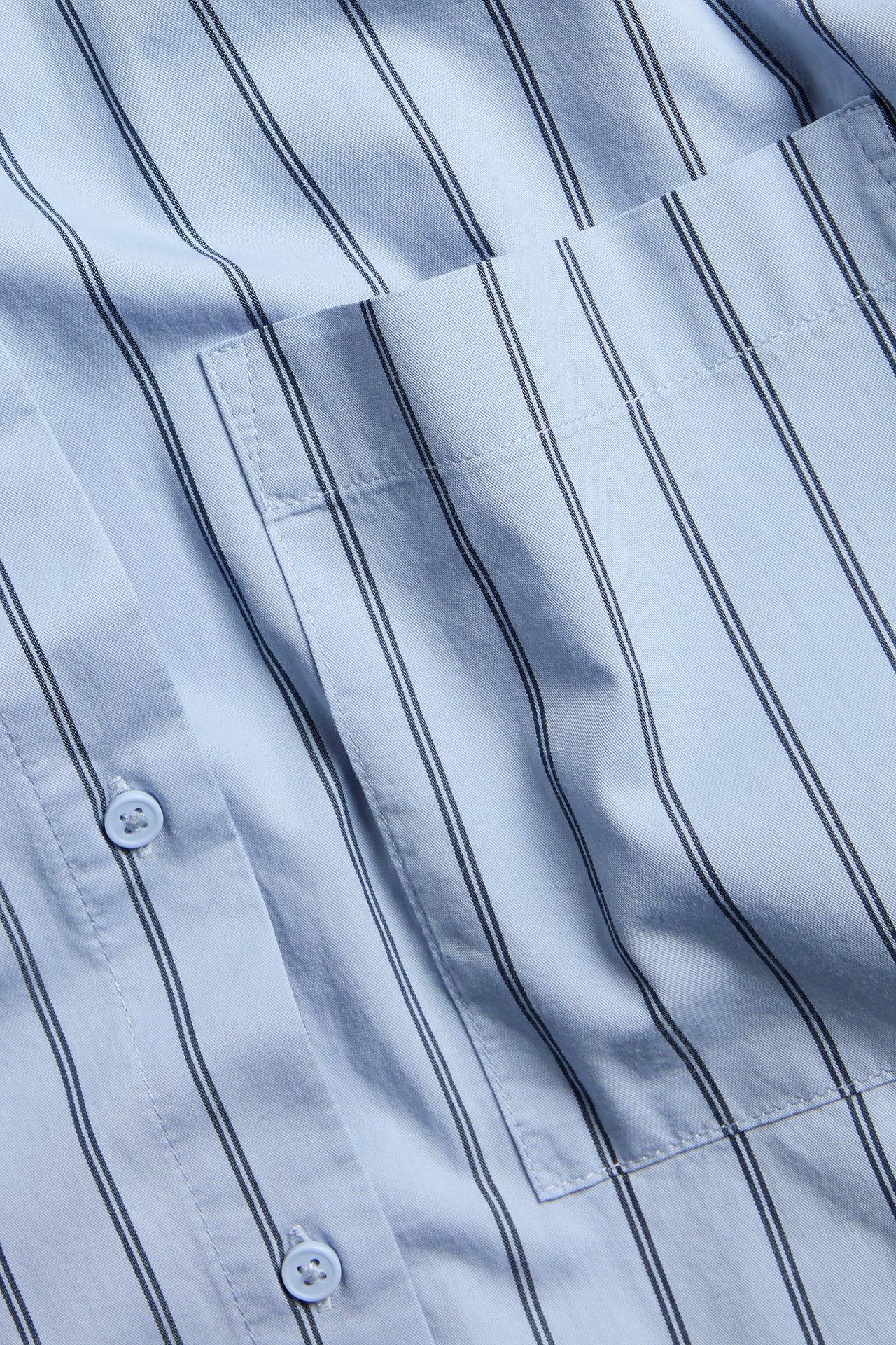 Zbliżenie na jasnoniebieską koszulę popelinową w paski z kieszenią na piersi i jasnoniebieskimi guzikami.