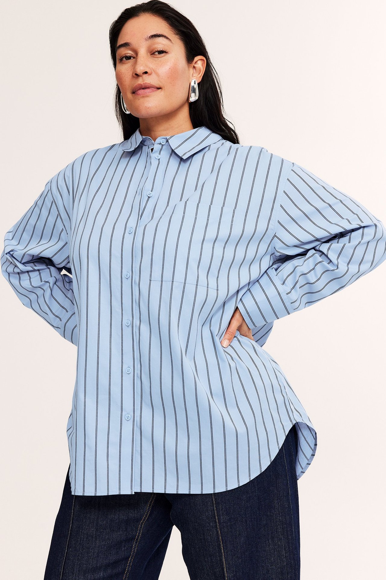 Widok z przodu: kobieta w oversize'owej jasnoniebieskiej koszuli w paski, ciemnoniebieskich dżinsach i srebrnych kolczykach.