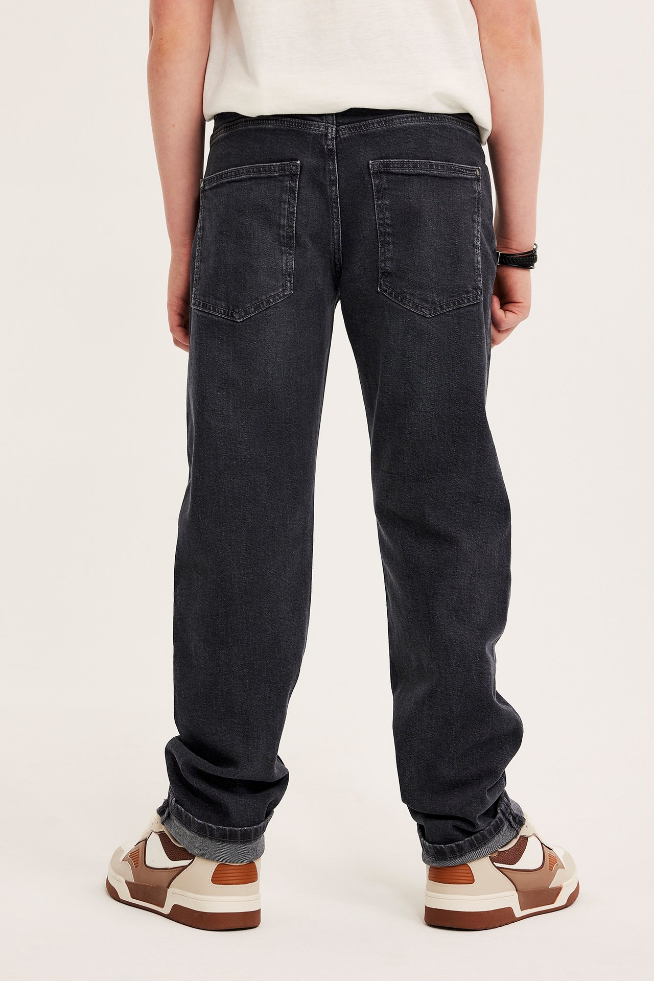 Bakfra: et barn i grå rette stretchjeans, hvit t-skjorte, brune joggesko, svart armbånd.