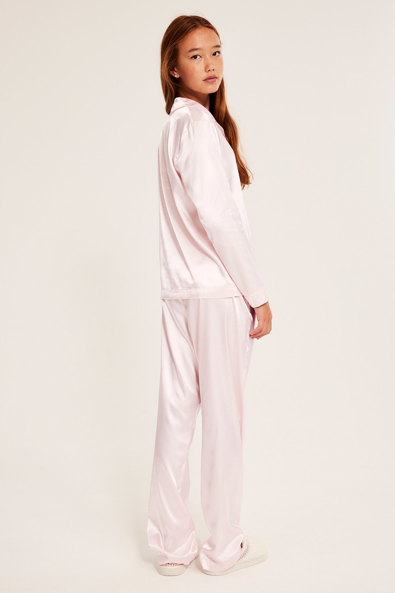 Sidovy av ett barn i ljusrosa pyjamas i satin med långa ärmar och ljusa tofflor.