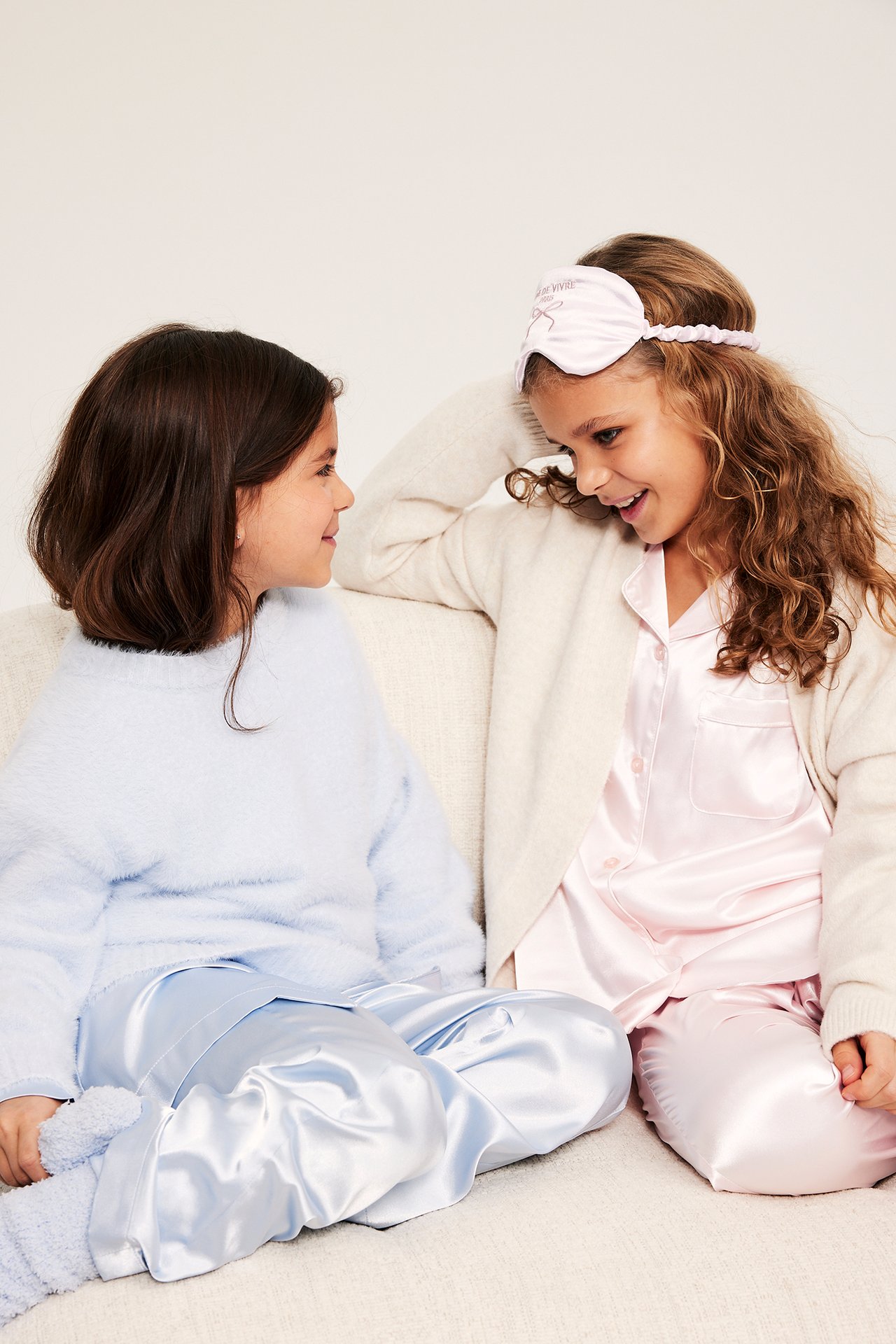 Sidovy av barn i ljusrosa satinpyjamas, ögonmask, kofta. Ett annat barn i ljusblå pyjamas, fluffig tröja.