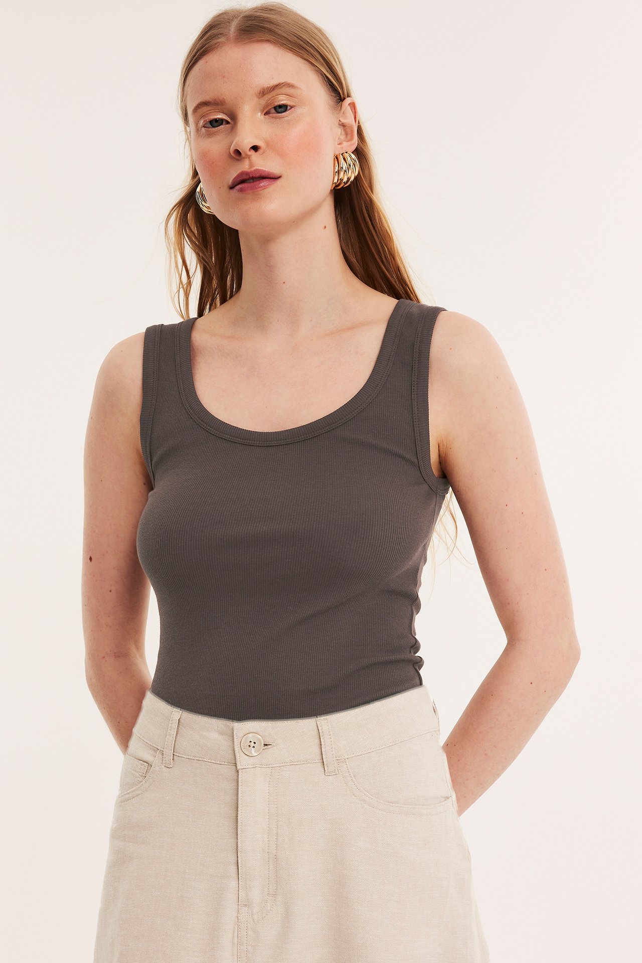 Framifrån: kvinna i mörkbrun ribbad tanktopp, ljusbeige shorts och guldfärgade ringörhängen.