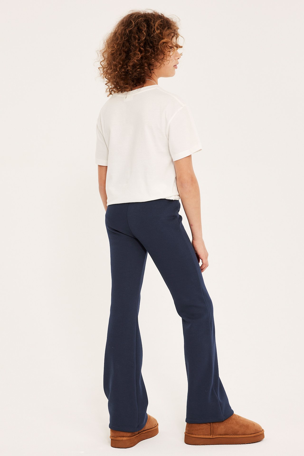 Lapsi takaa päin tummansinisissä levenevissä leggingseissä, valkoisessa t-paidassa ja ruskeissa saappaissa.