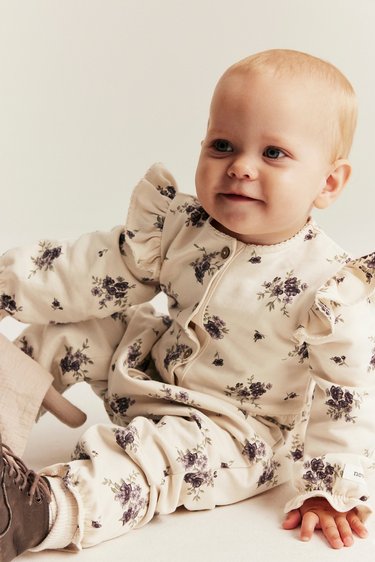 Frontvisning av en baby i lys beige blomstret bomullsjumpsuit med skulderkapper og knapper foran.