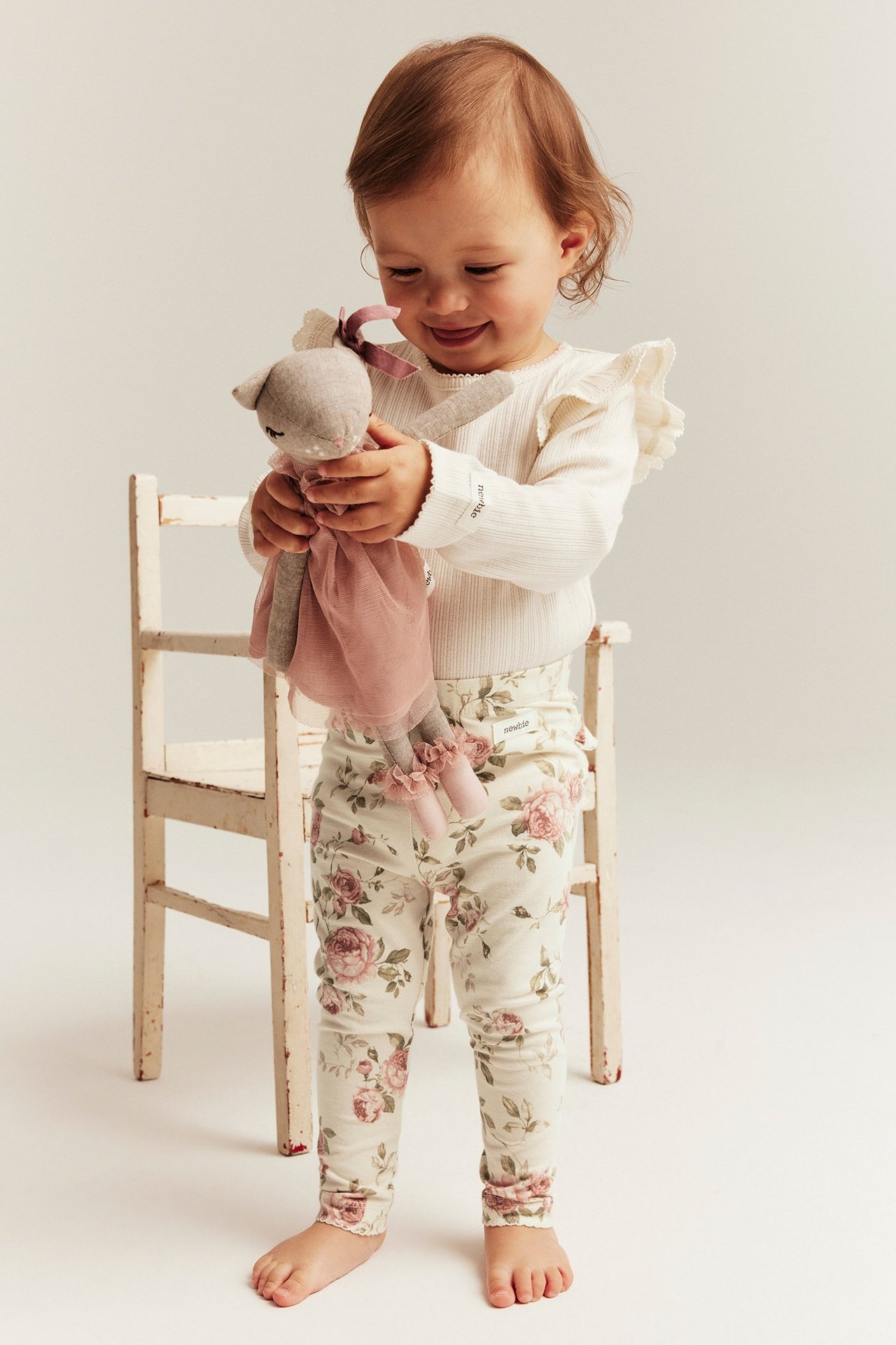 Edestäpäin vauva yllään vaaleanbeiget kukkakuosiset leggingsit vaaleanpunaisilla ruusuilla ja valkoinen ribbitoppi, pitäen nukkea.