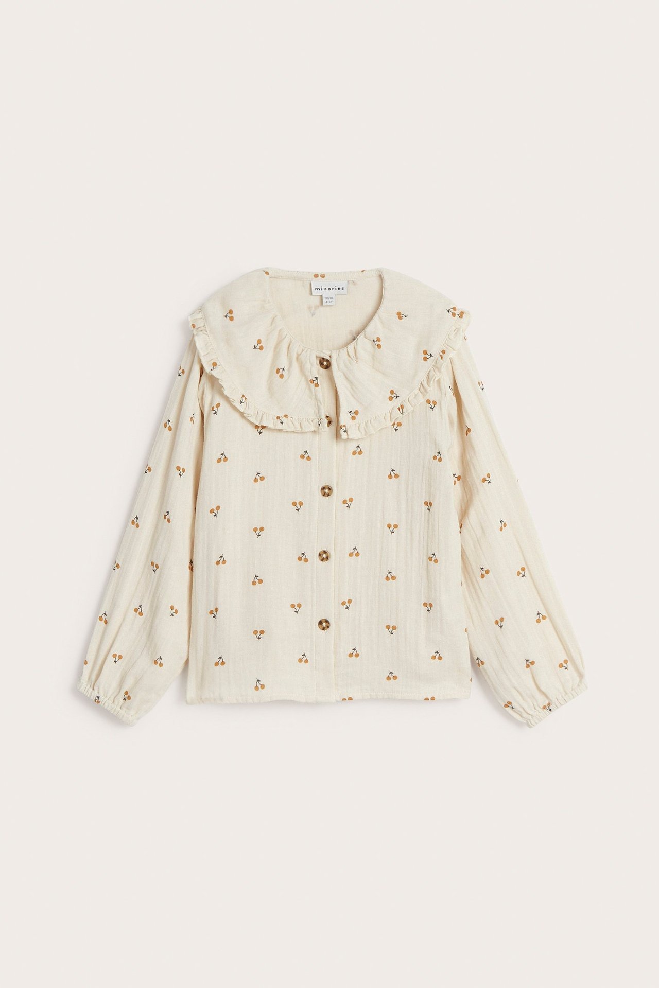 Framifrån: Barnblus i beige bomull med volangkrage, körsbärsmönster och knappar framtill.