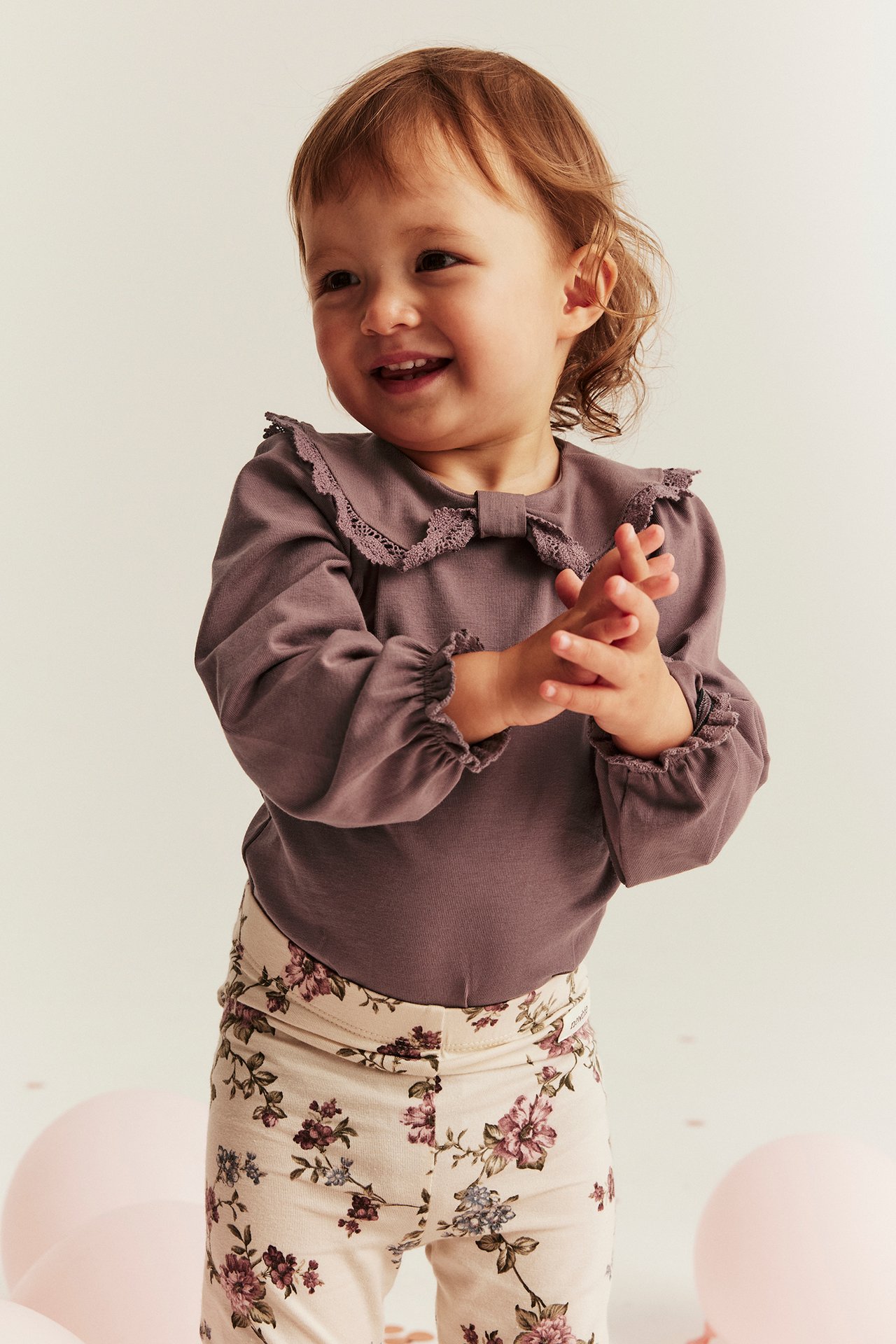 Framifrån: Leende baby i mörkrosa långärmad body med spetskrage, rosett och blommiga leggings.