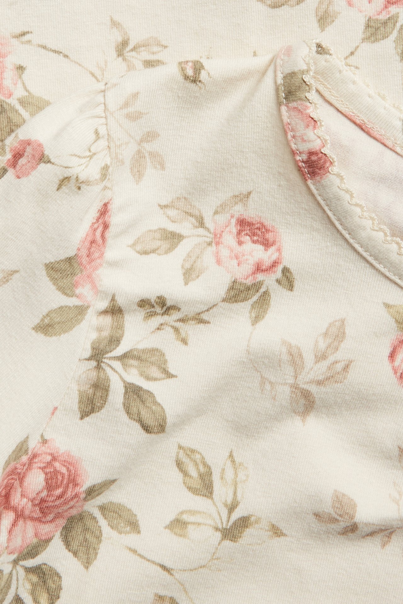 Nærbilde av en lys beige topp med rosa blomstermønster, puffermer og picotkant ved halsen.